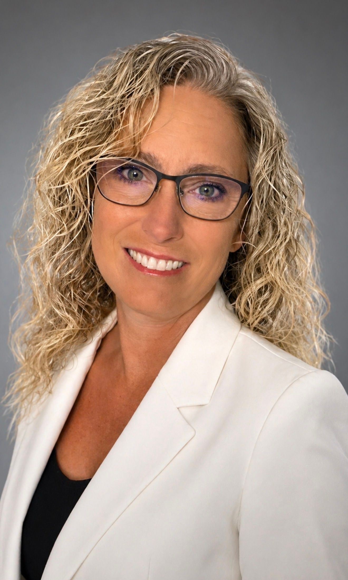 Professional woman with curly blonde hair and glasses wearing a white blazer smiling confidently.