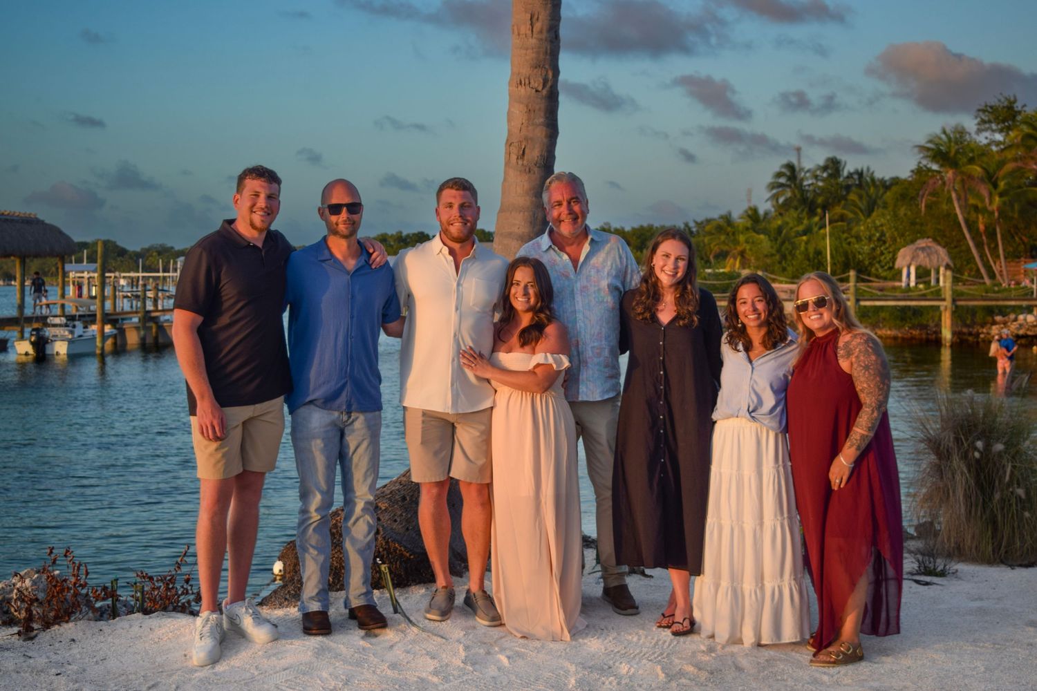 Group of eight adults posing together on a sandy beach at sunset near a tropical marina.