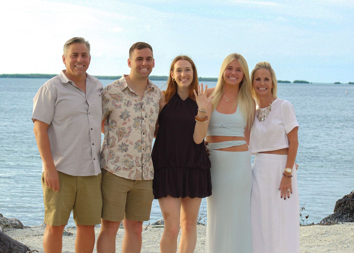 Five smiling people pose together on a rocky beach with calm ocean waters and a cloudy sky in the background.