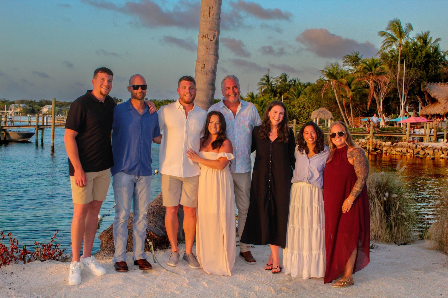 Group of seven friends posing together on a sandy beach at sunset near a tropical waterfront.