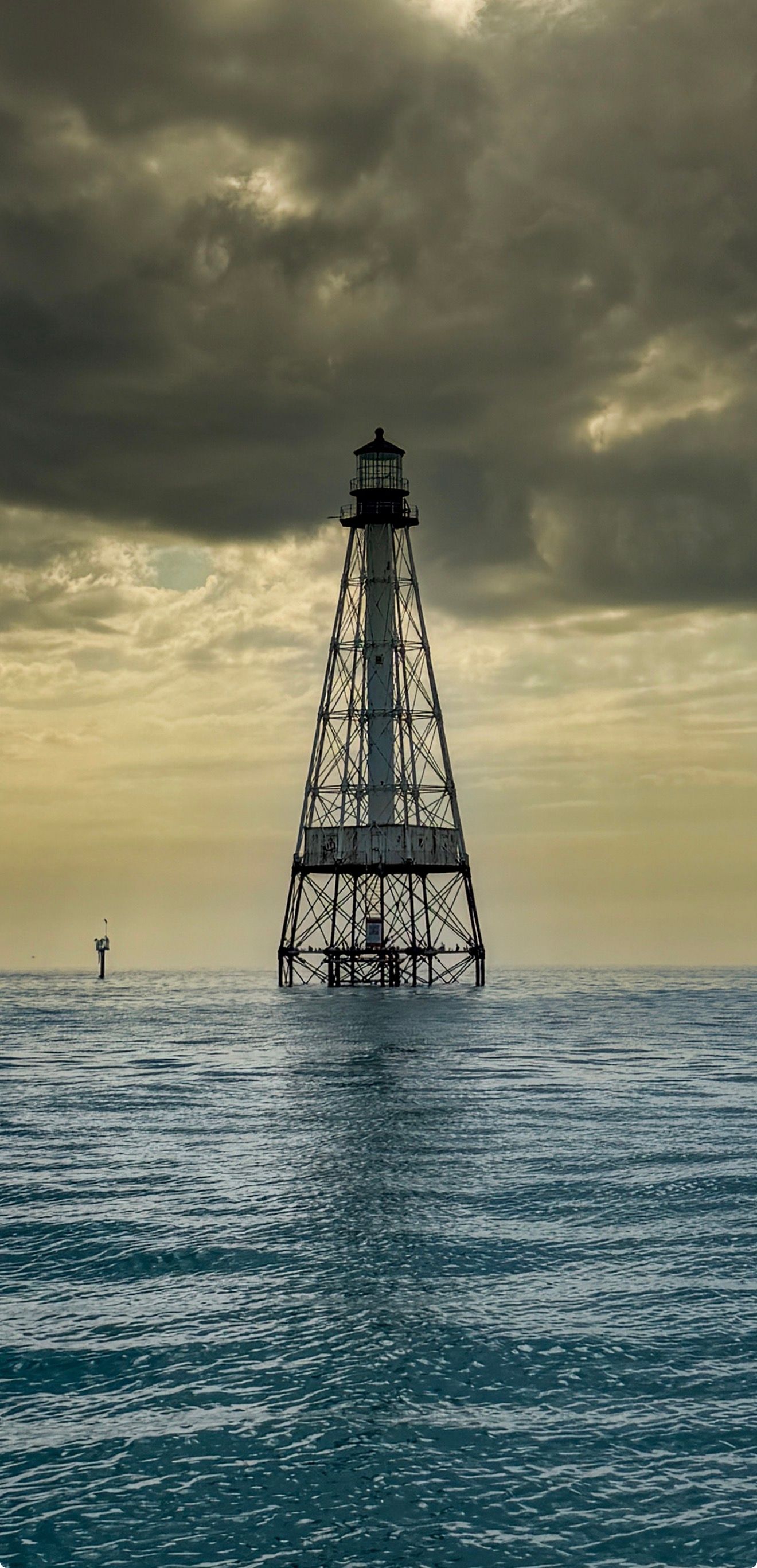 A solitary offshore lighthouse stands on metal stilts amid choppy blue ocean waters under dramatic storm clouds.