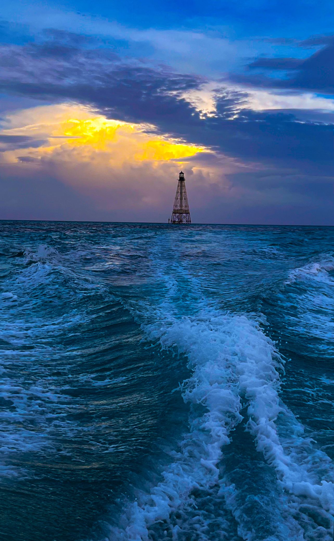 A lighthouse stands in choppy turquoise ocean waves under a dramatic sunset sky with golden clouds.