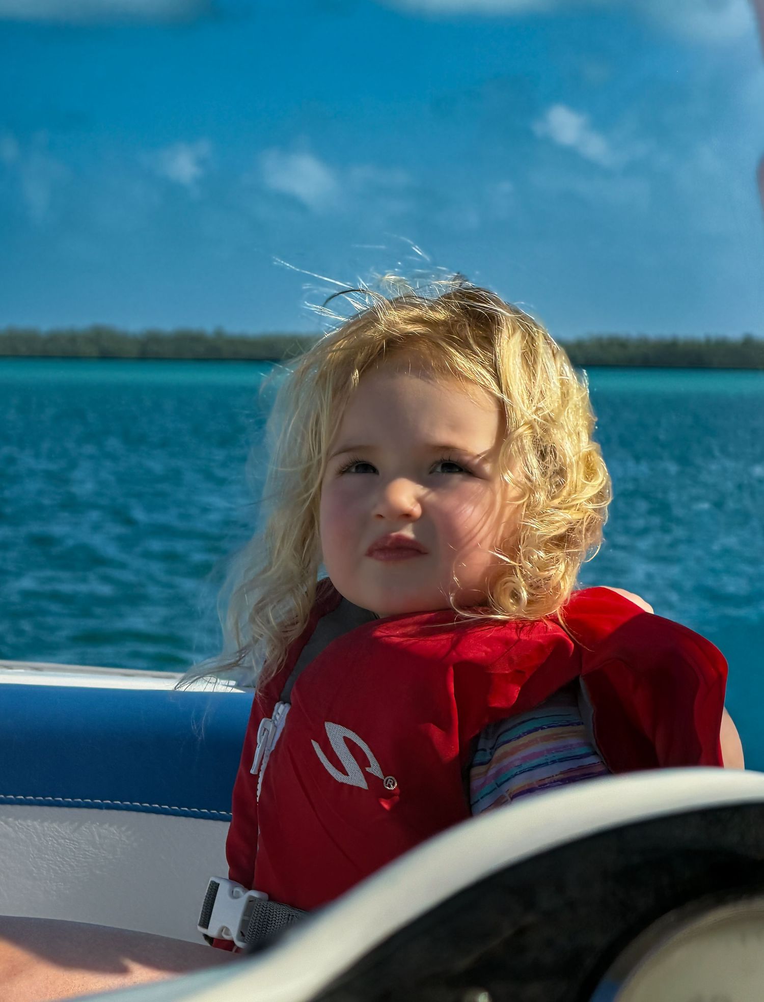 Young blonde child wearing a red life jacket on a boat with turquoise ocean water and blue sky in background.