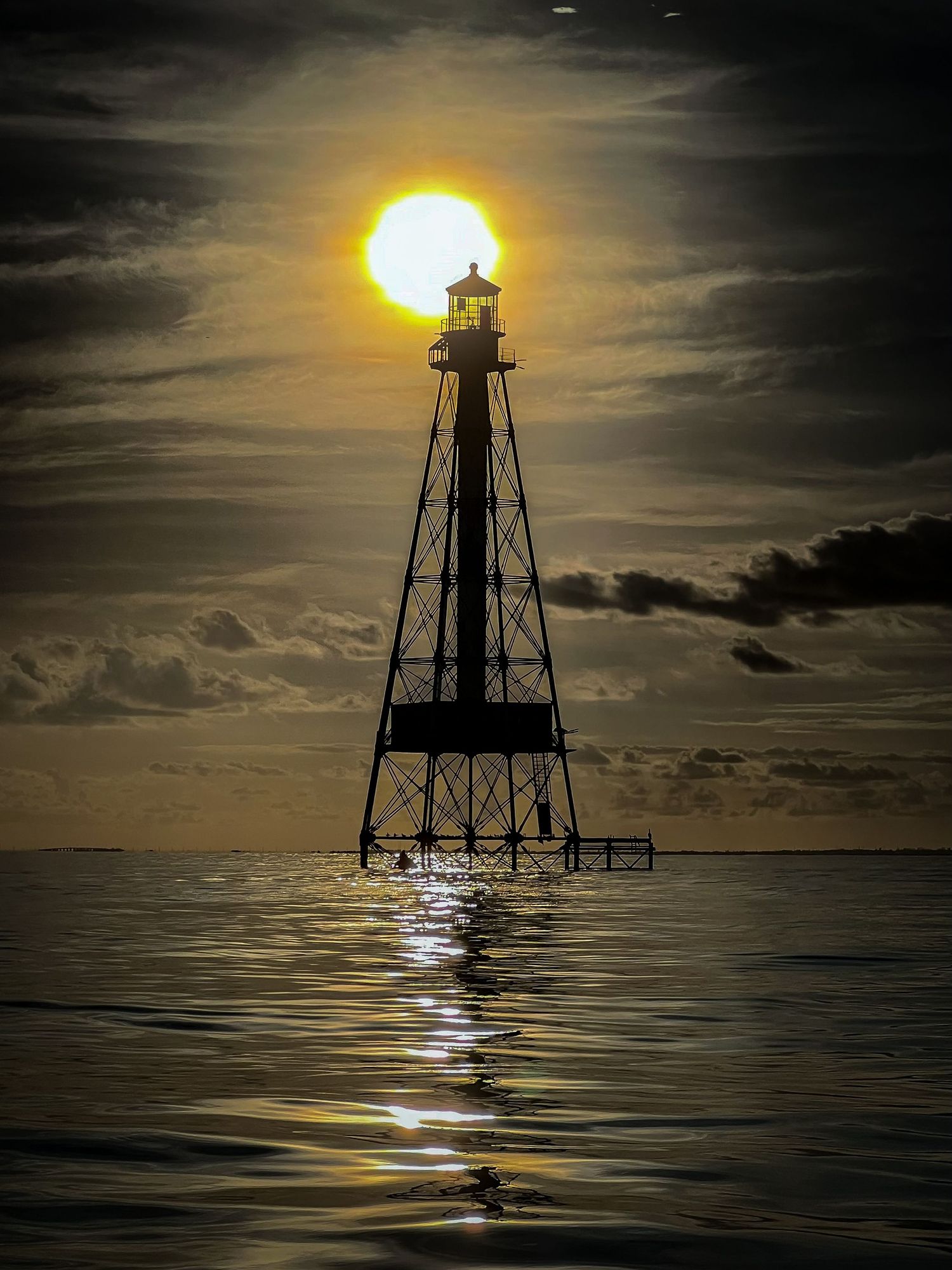 Dramatic lighthouse silhouette against glowing golden sun reflecting over dark ocean waters.