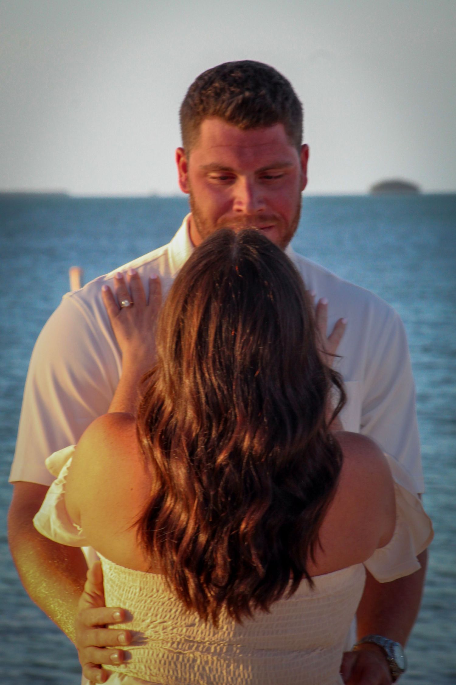 Couple embracing at sunset by the ocean, man in white shirt holding woman with dark wavy hair near a seaside pier.