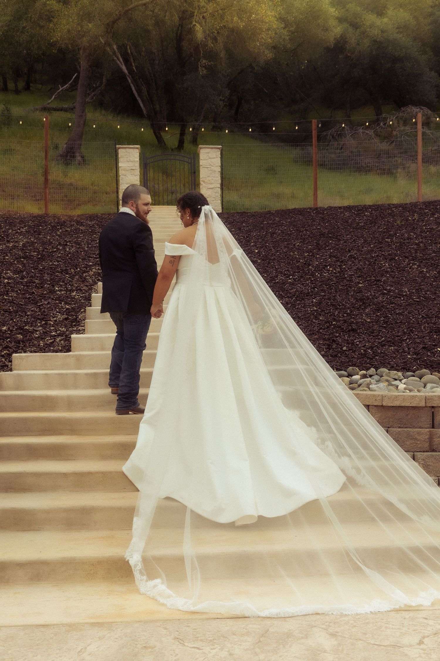 Bride in flowing white gown and long veil walks with groom up stone steps at outdoor wedding venue.