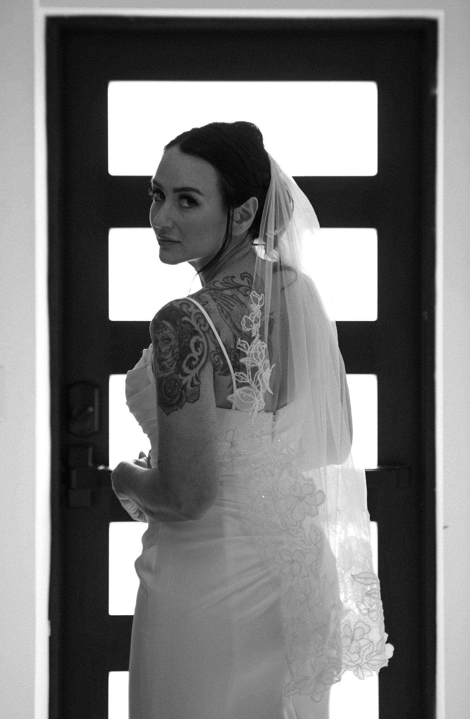 Black and white photo of a tattooed bride in a lace wedding dress and veil, looking over her shoulder by a door.