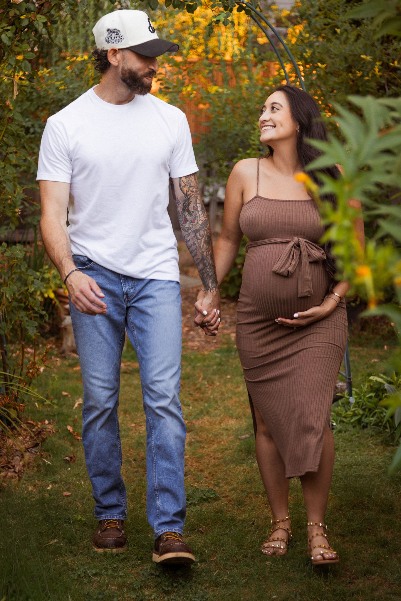 Couple walking through autumn foliage wearing brown and white outfits during maternity photoshoot.