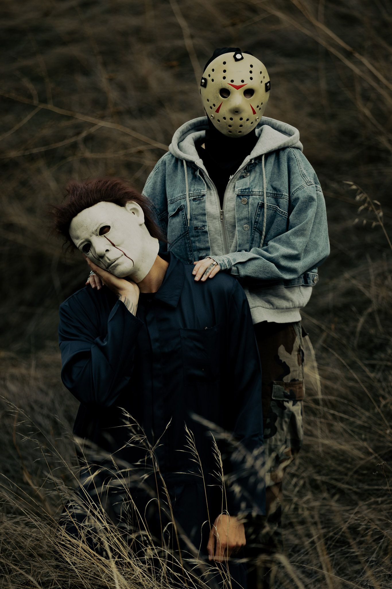 Two people wearing horror movie masks pose dramatically in tall grass at dusk.