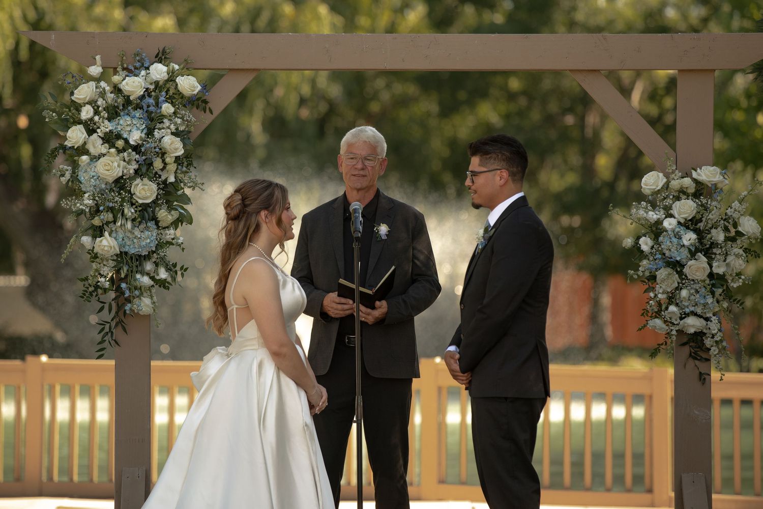 Bride and groom exchange vows under a wooden arch adorned with white and blue floral arrangements at an outdoor wedding ceremony.