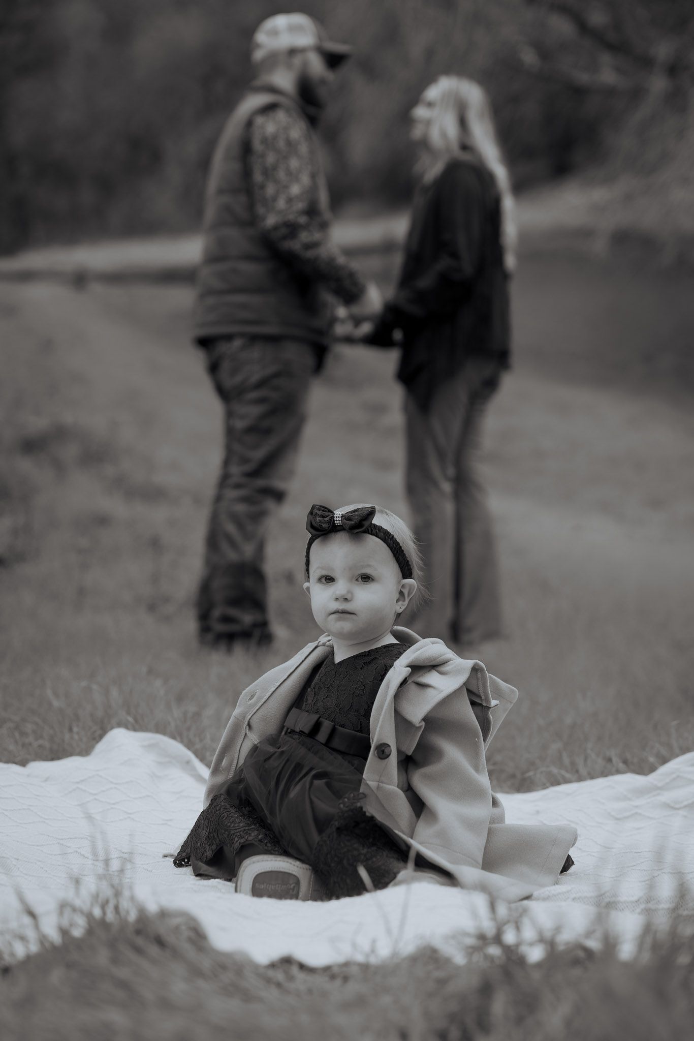 A baby in a dress sits on a blanket in the foreground while two people converse in the background in black and white.