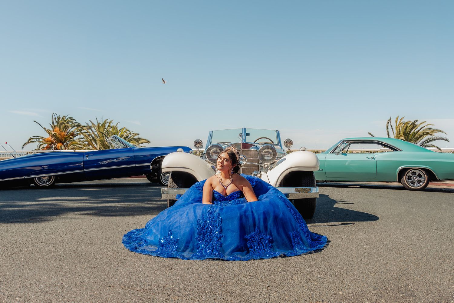 A blue evening gown flows dramatically across a parking lot with palm trees and cars in the background.