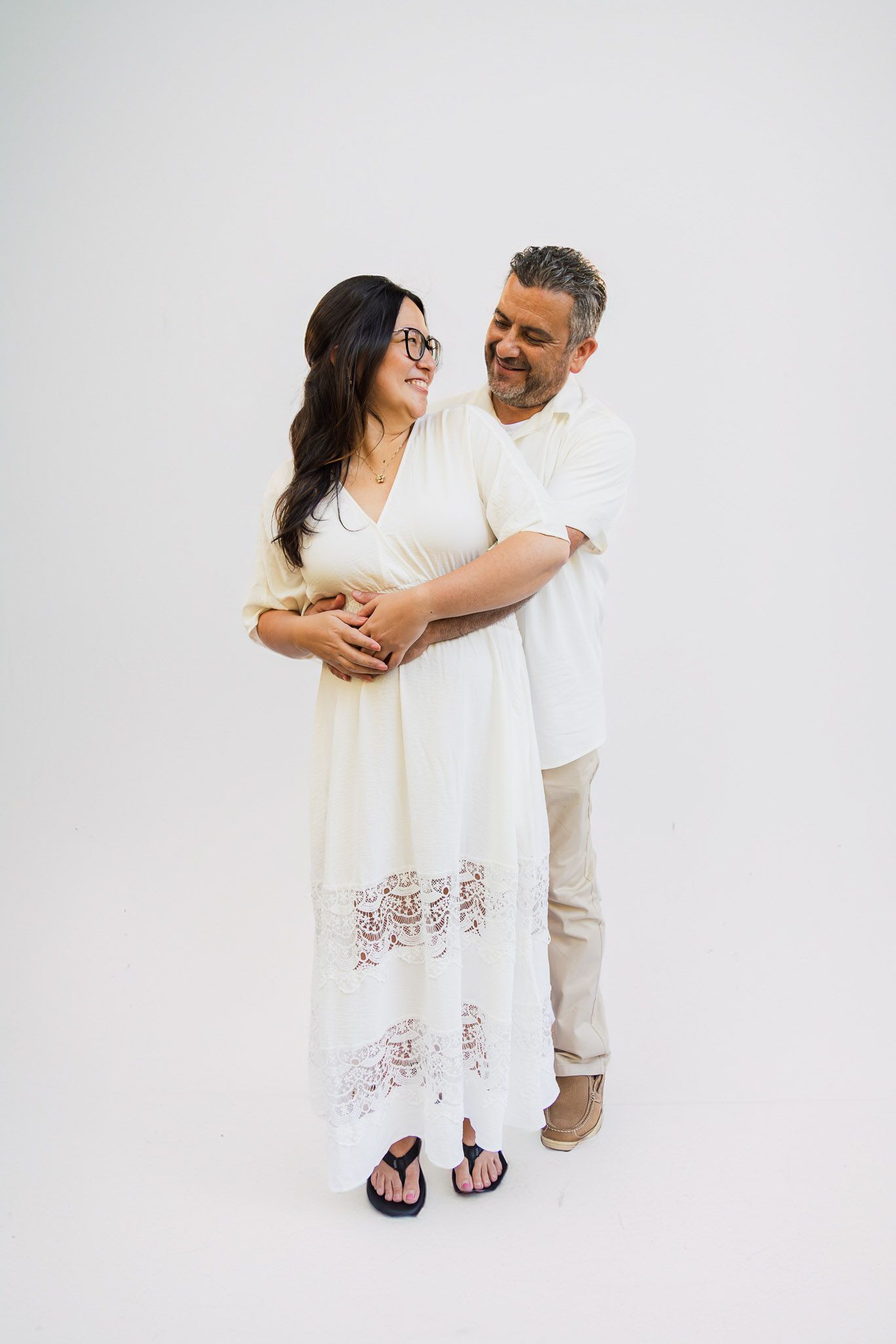 A couple wearing white outfits embraces and smiles together against a light background in a romantic photoshoot.