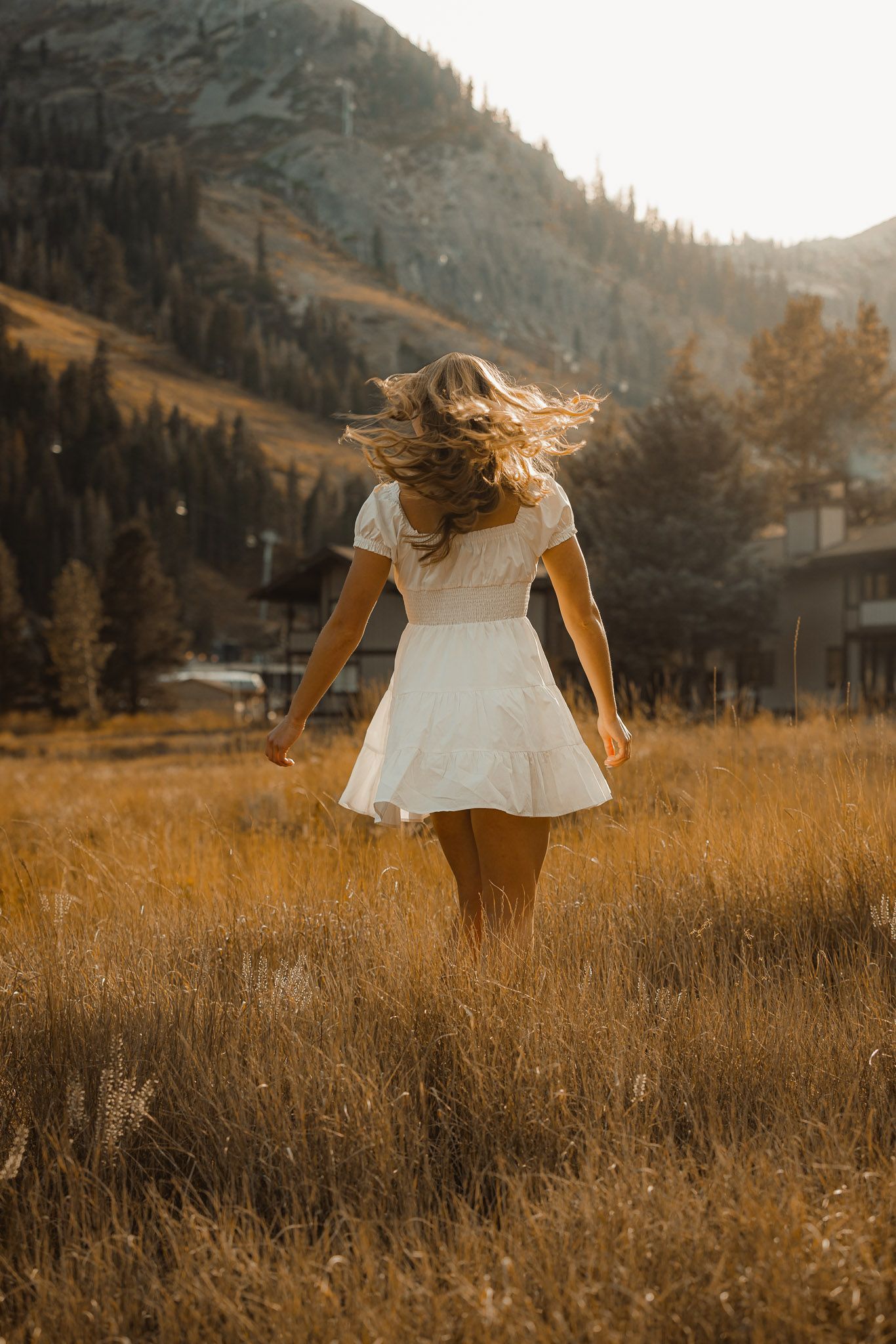 A figure in a white dress stands in a golden autumn field with mountains in the background during sunset.