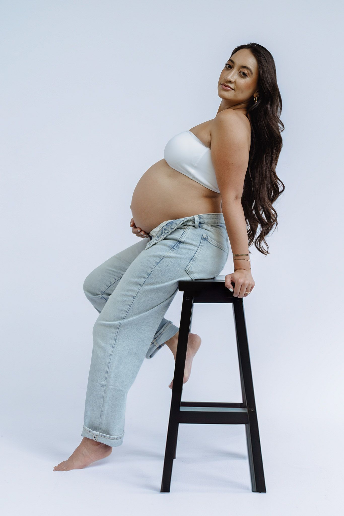 Artistic maternity studio portraits showing pregnant woman in casual attire posing on black stool.