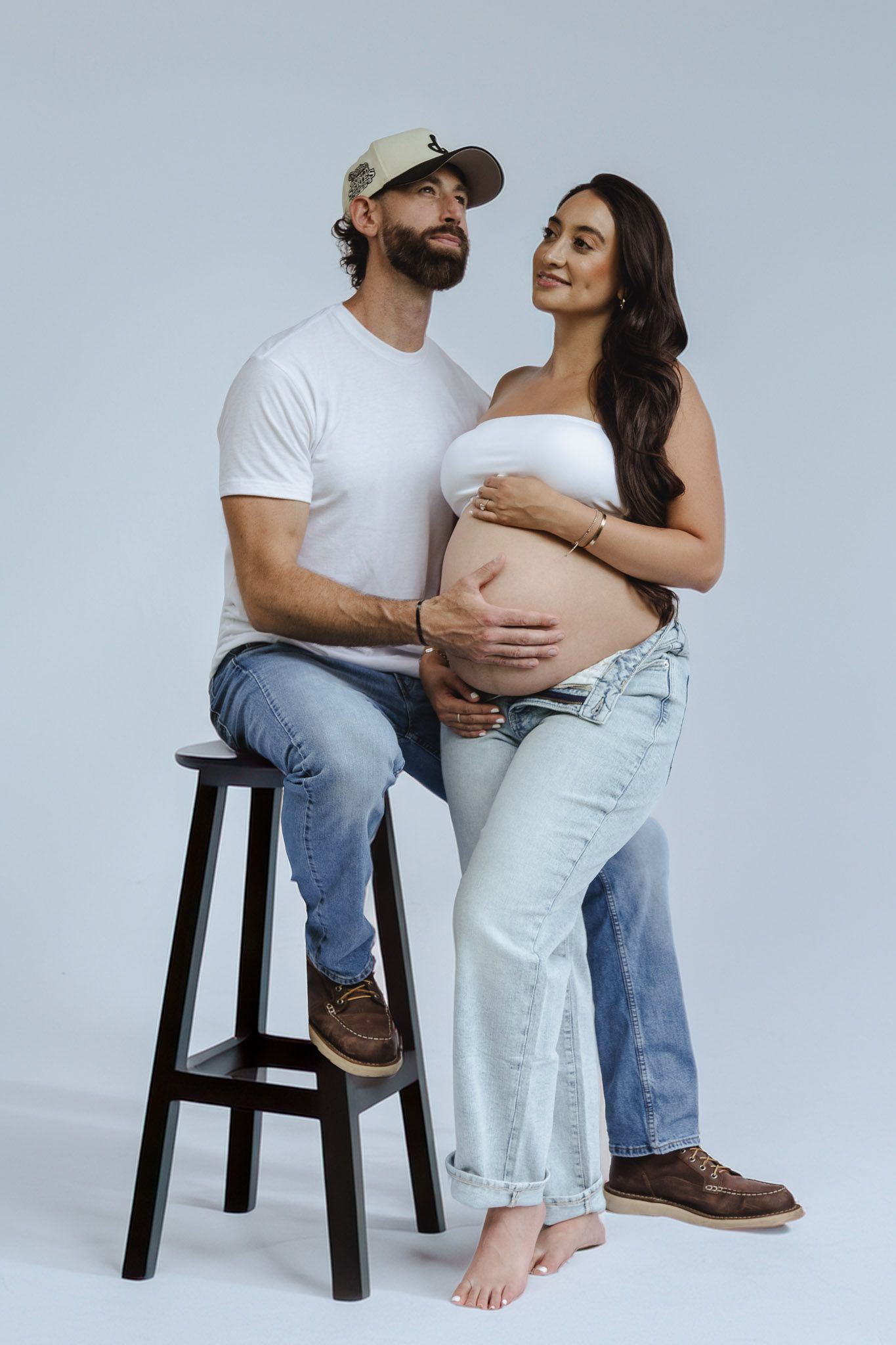Clean studio maternity portraits featuring couple in white and denim against light background.