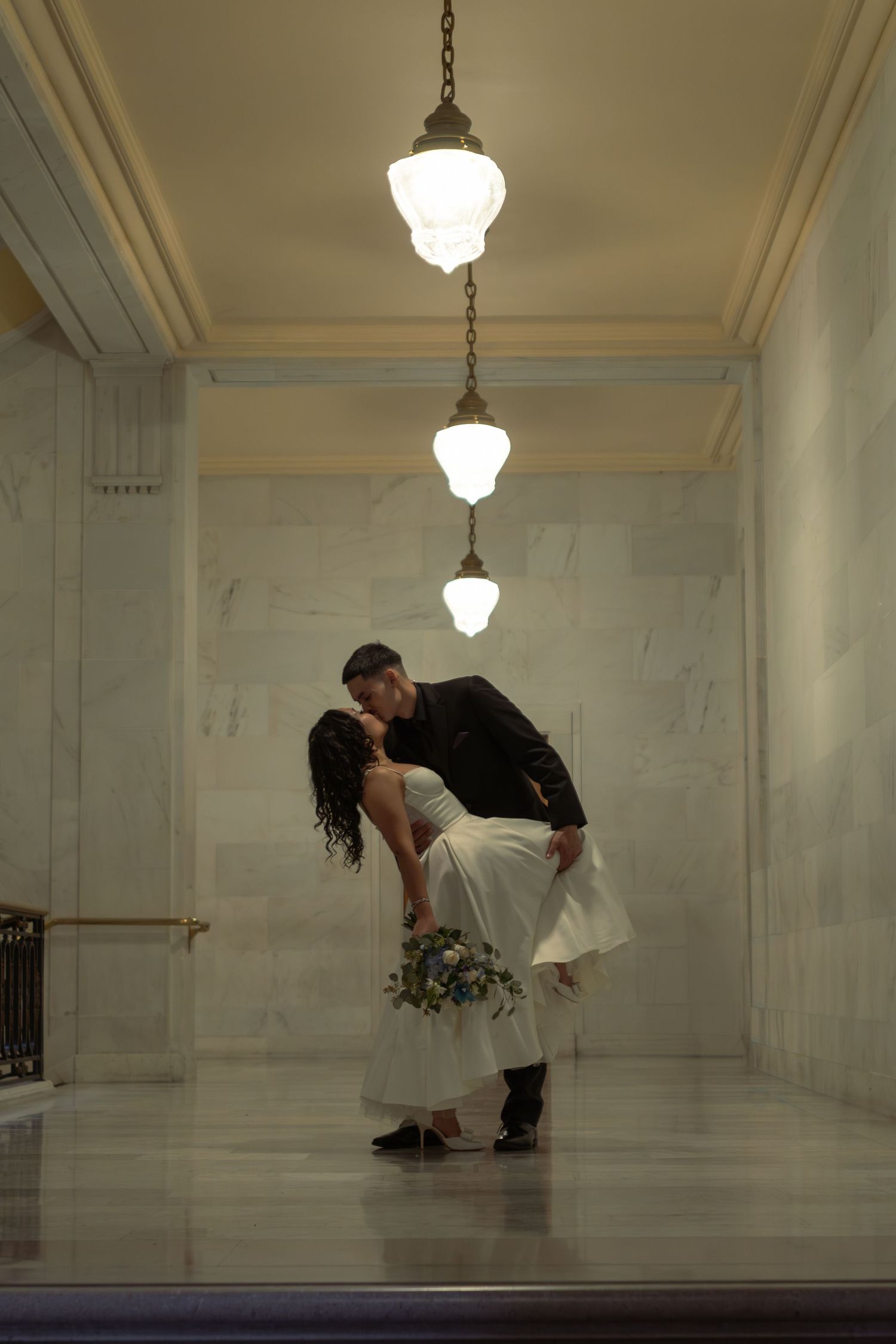 Couple sharing a romantic dip kiss in an elegant marble hall, bride in white gown holding bouquet, groom in black suit.