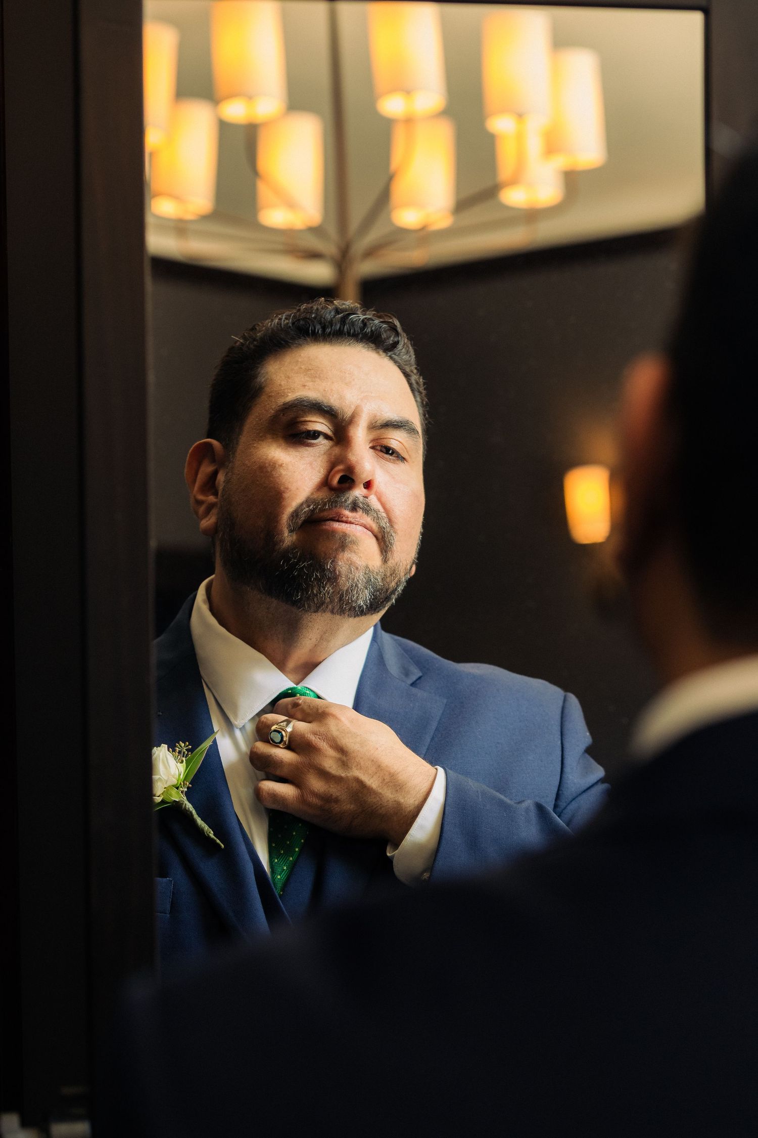 Groom in blue suit adjusting green tie in mirror reflection before wedding ceremony.