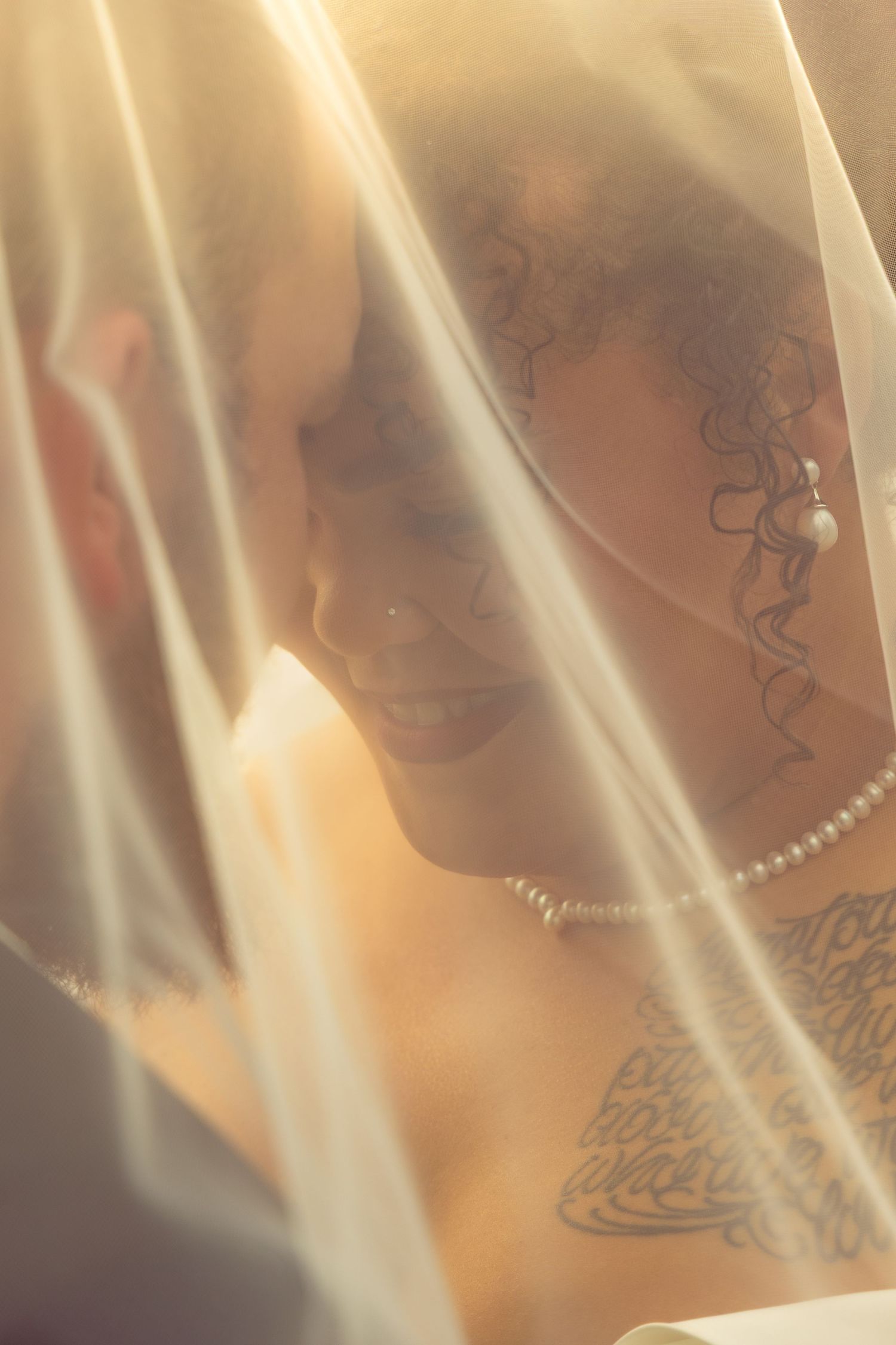 Bride and groom share an intimate moment under a sheer wedding veil, bride wearing pearl jewelry and tattoos.