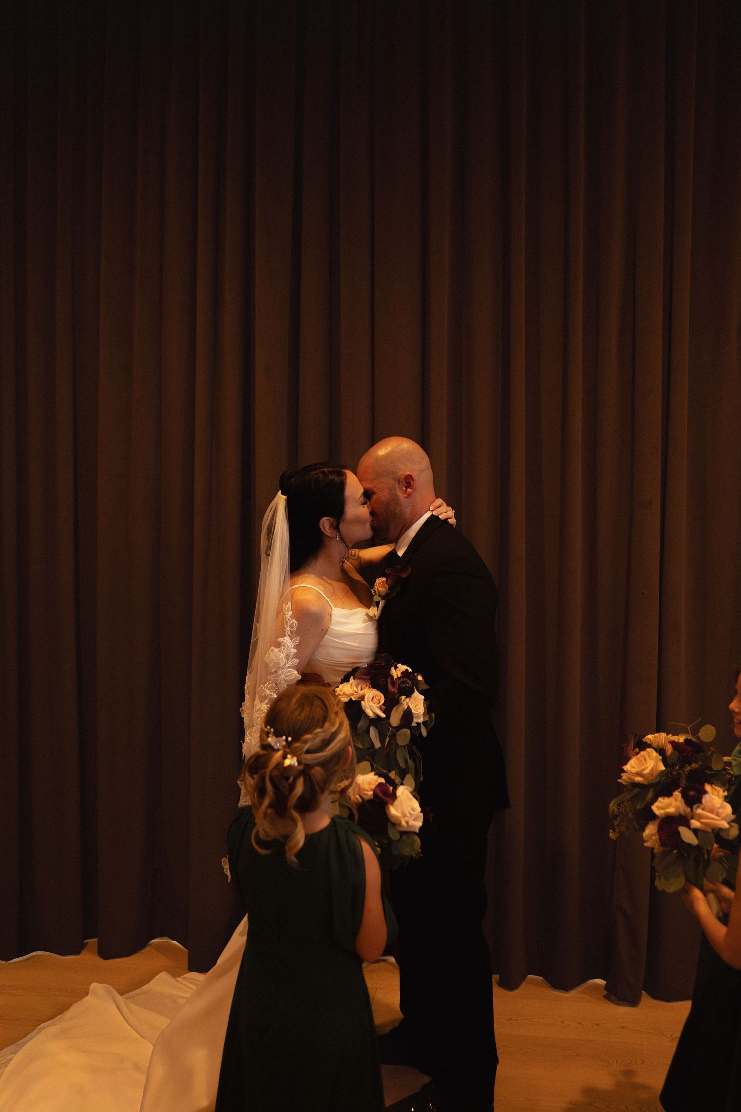 Bride and groom share first kiss at wedding altar, bride in white gown with veil, groom in black tuxedo.