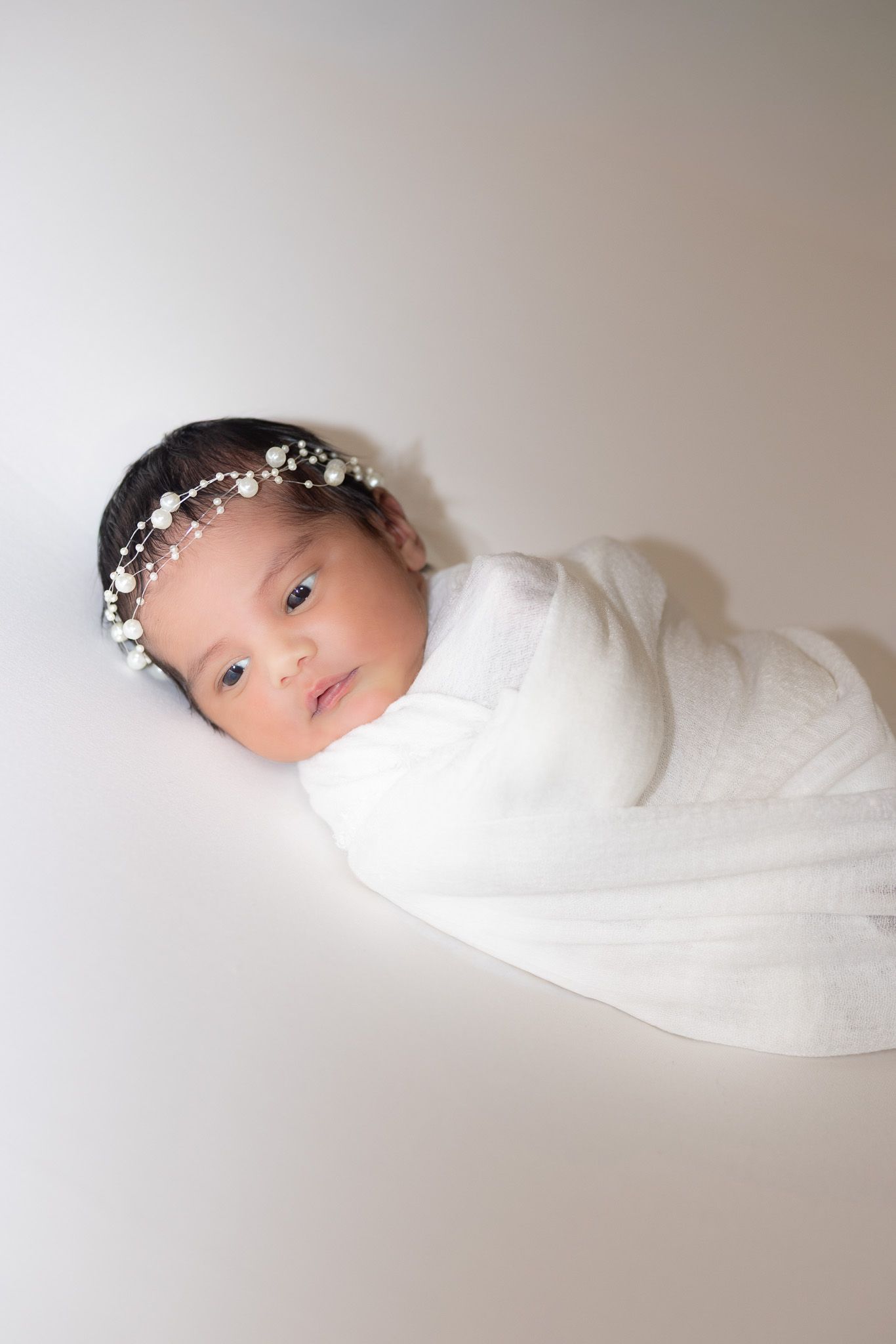 A newborn baby swaddled in white fabric wearing a delicate floral headpiece against a light background.