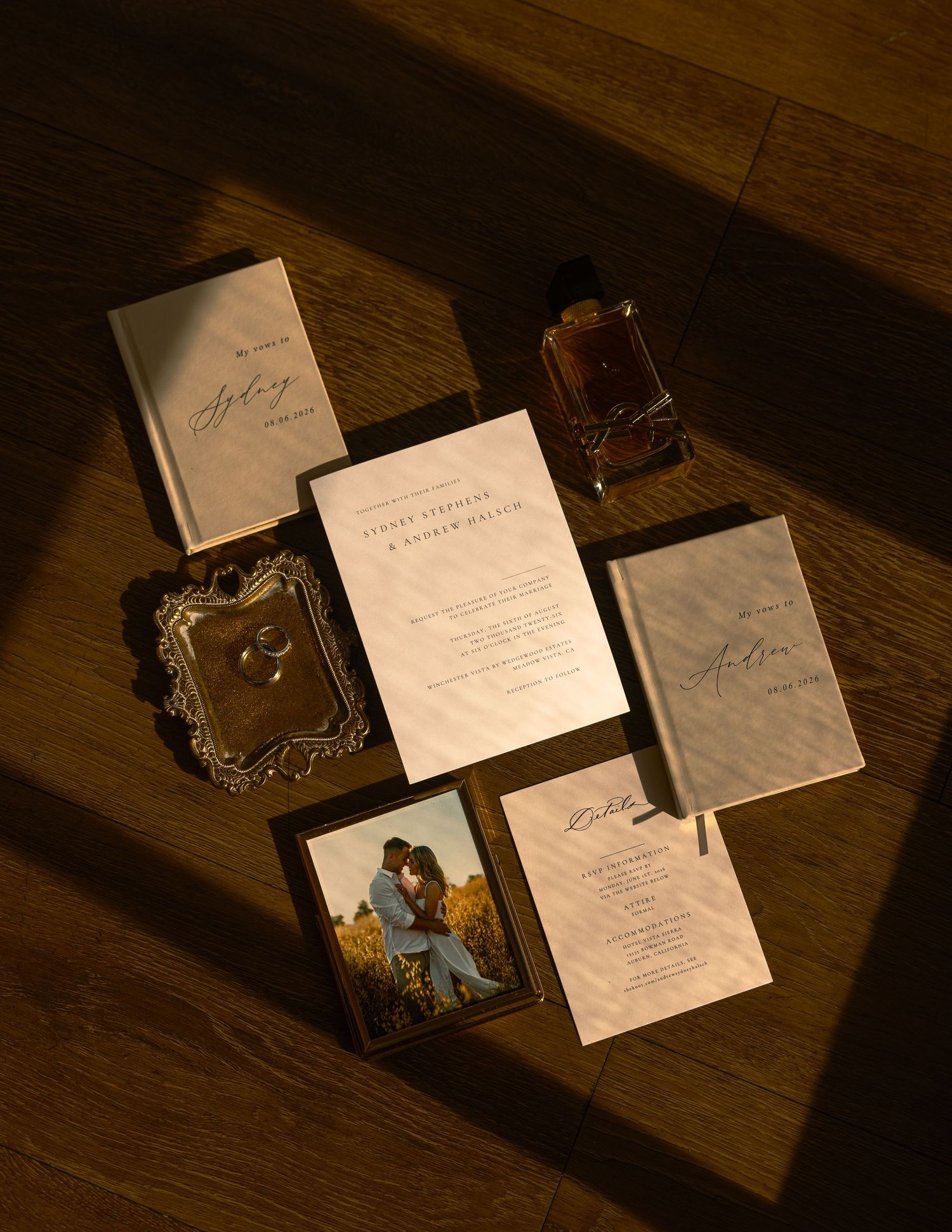 Elegant wedding invitation suite with stationery, rings, couple photo, and perfume bottle on warm wooden floor.