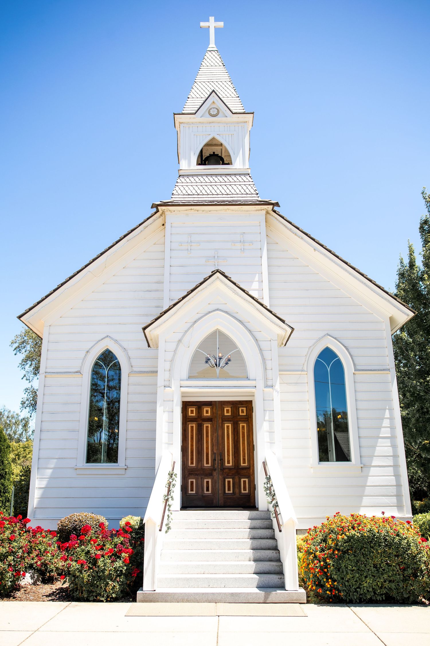 White wooden chapel with steeple, cross, arched windows, and wooden doors surrounded by colorful flowers under blue sky.