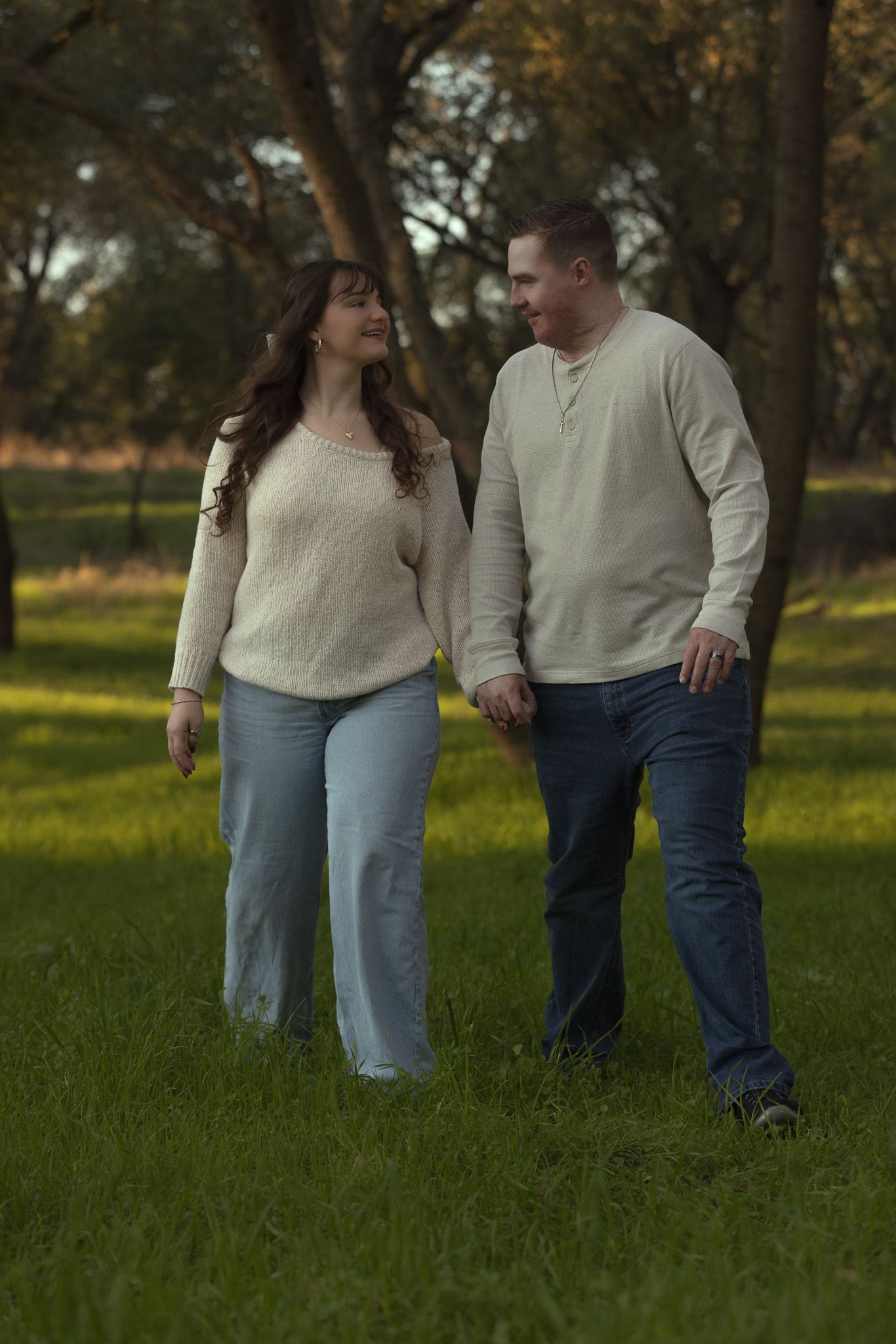 A couple in matching cream sweaters and jeans walks together through a sunlit grassy field.