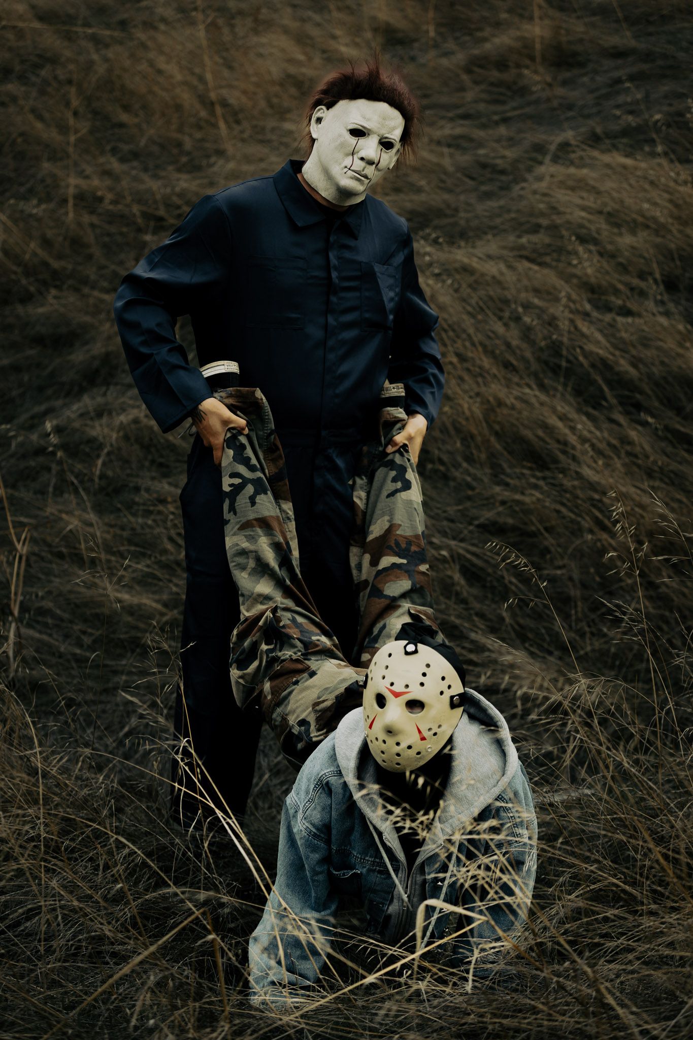 Two horror movie masks shown in dark moody lighting against dried grass background.