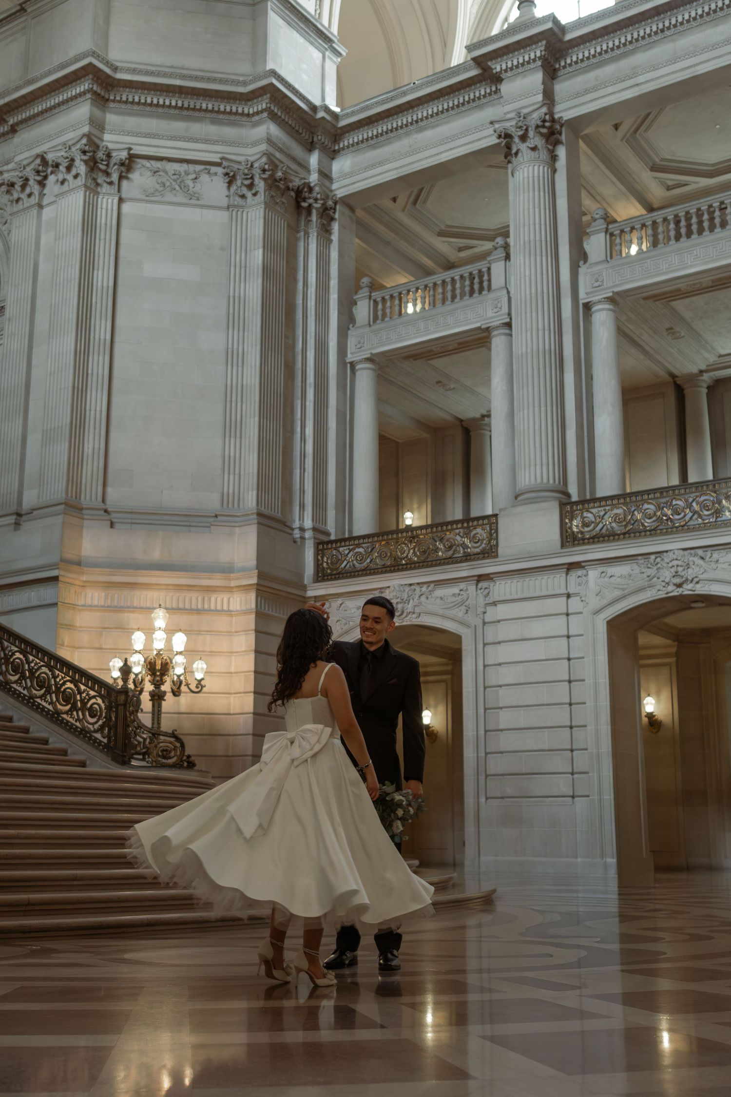 Bride in white dress and groom share romantic moment in grand marble hall with ornate staircase.