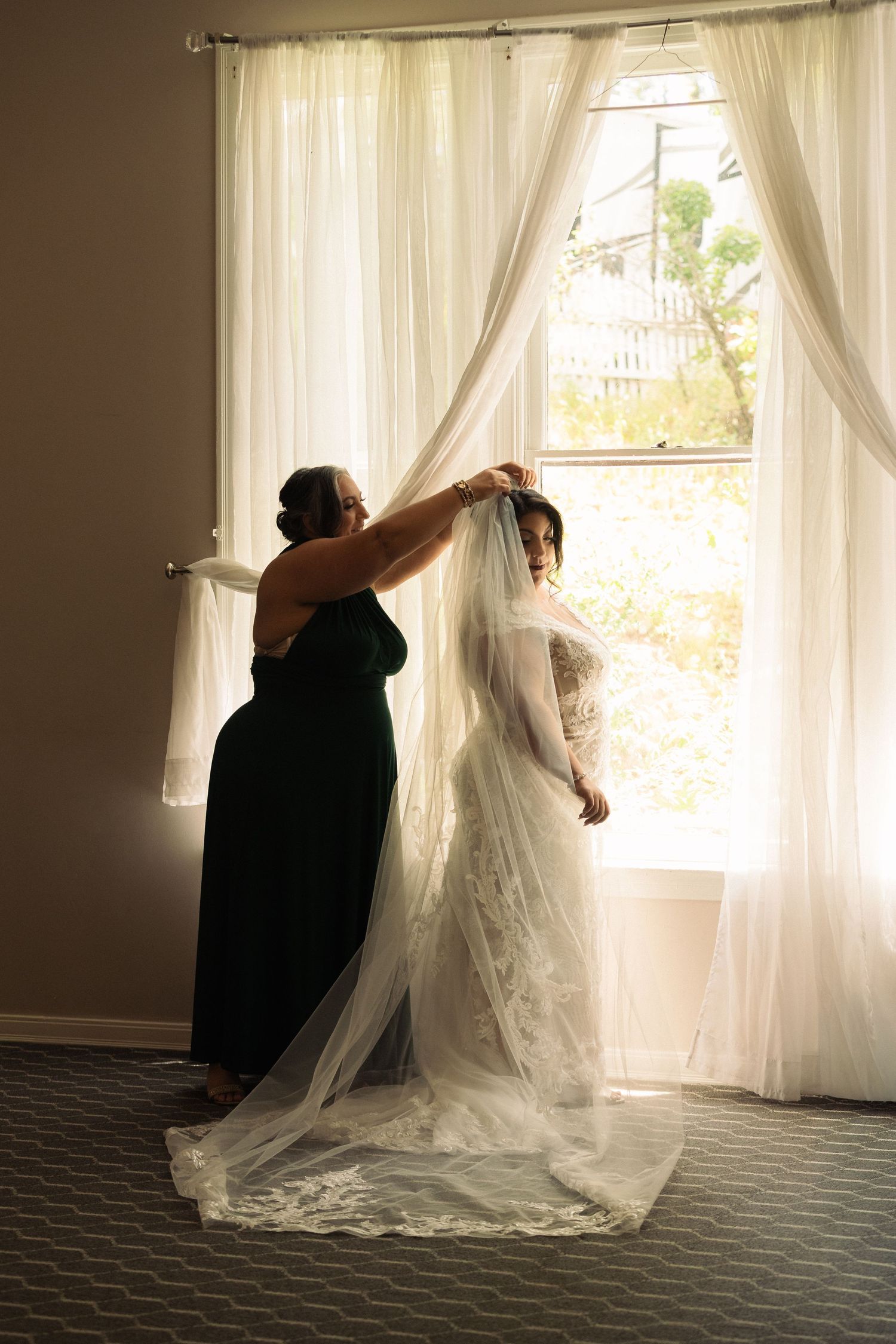 Bridesmaid in green dress helps bride with veil near sunlit window before wedding ceremony.