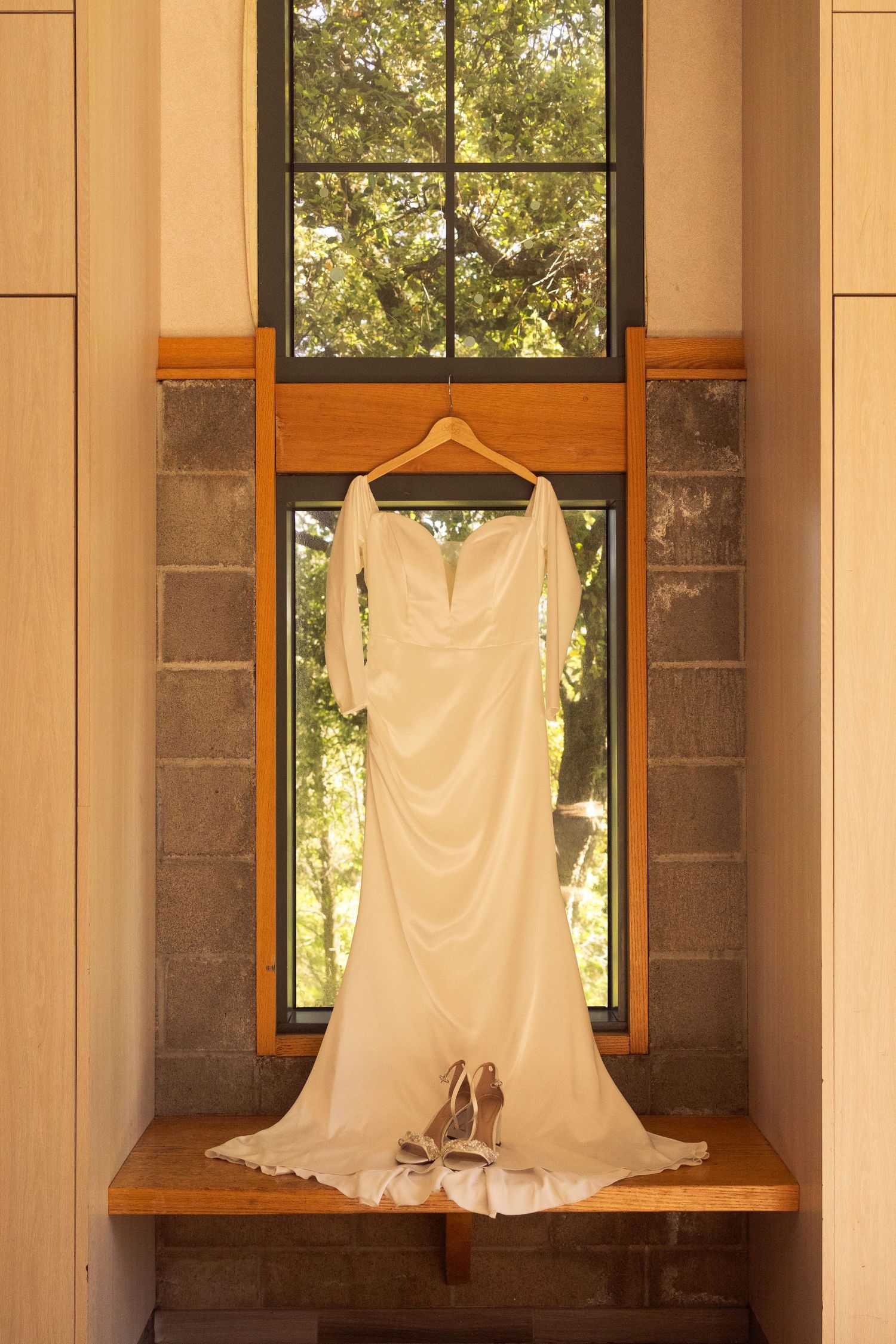 Elegant white wedding dress hanging in a window niche with heels below, natural light and trees visible outside.