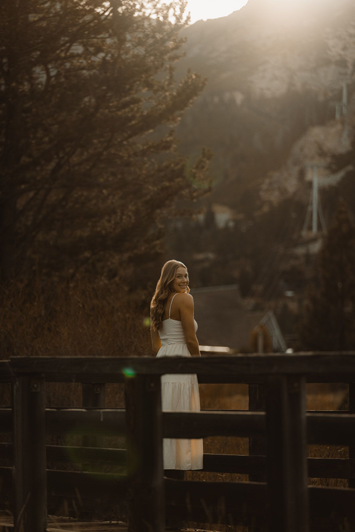 A woman in a white dress leans against a wooden fence at sunset in a dreamy outdoor setting.