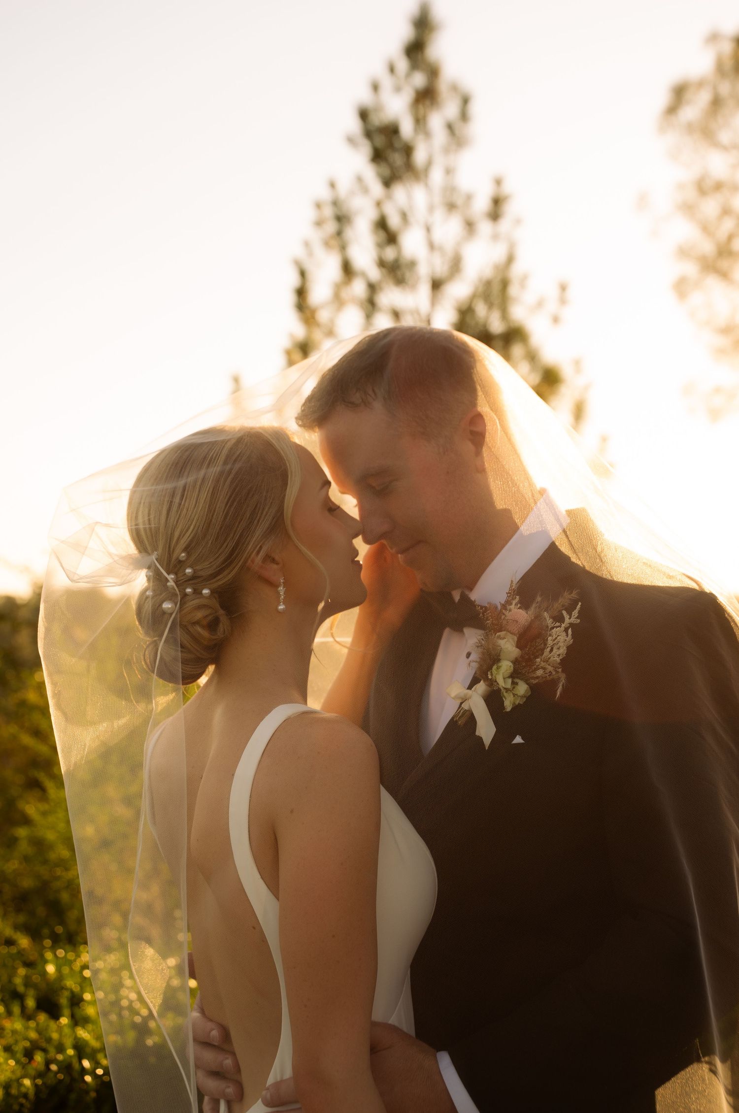 Bride and groom sharing romantic moment under veil in golden sunset light, surrounded by lush green trees.