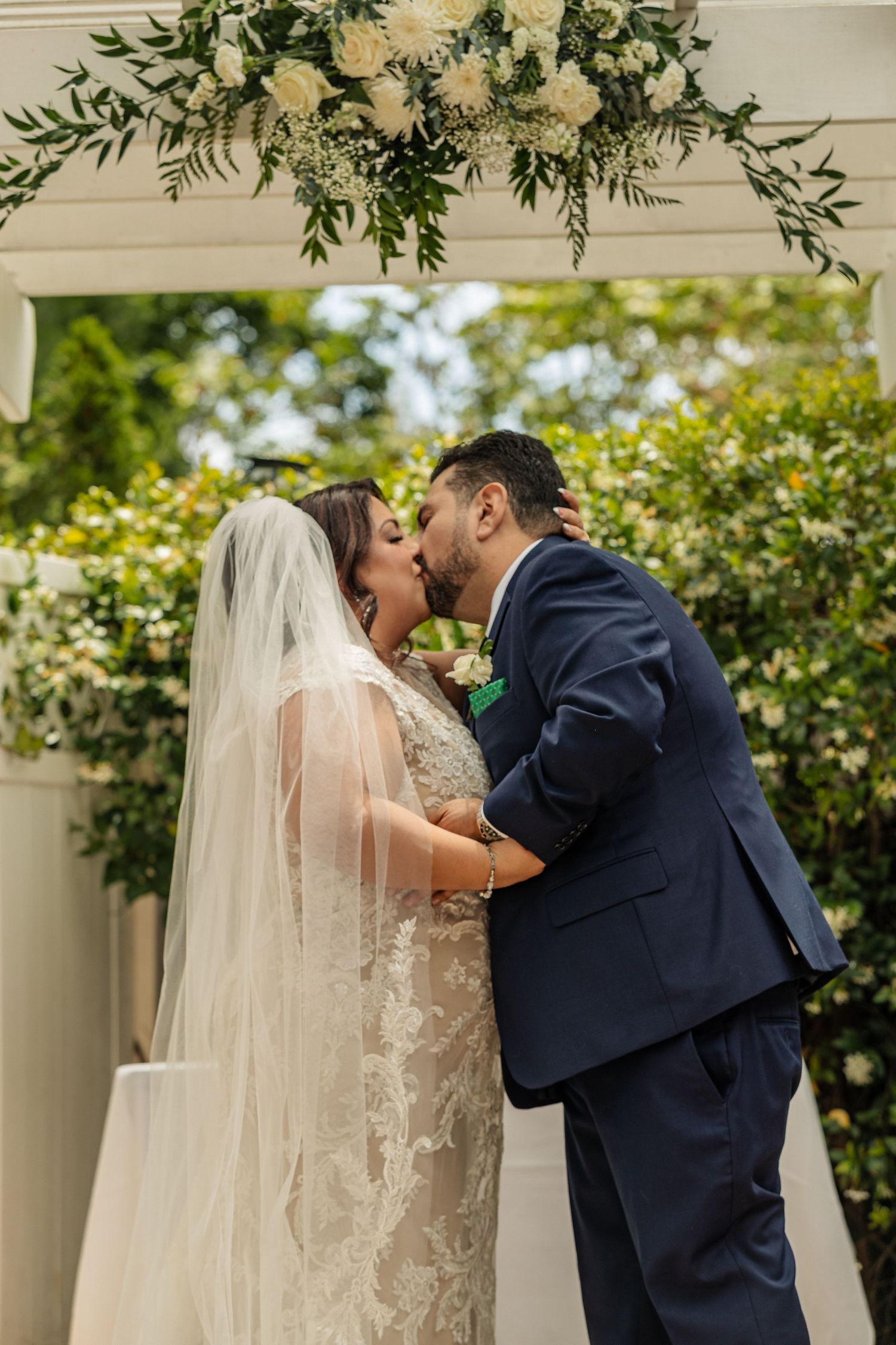 Bride and groom share their first kiss under a floral arch with white roses and greenery at an outdoor wedding ceremony.
