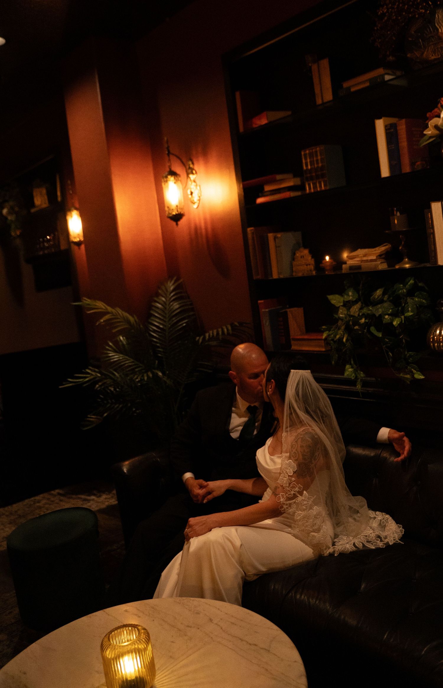 Bride and groom sit together on a leather sofa in a warmly lit, cozy library lounge during their wedding reception.