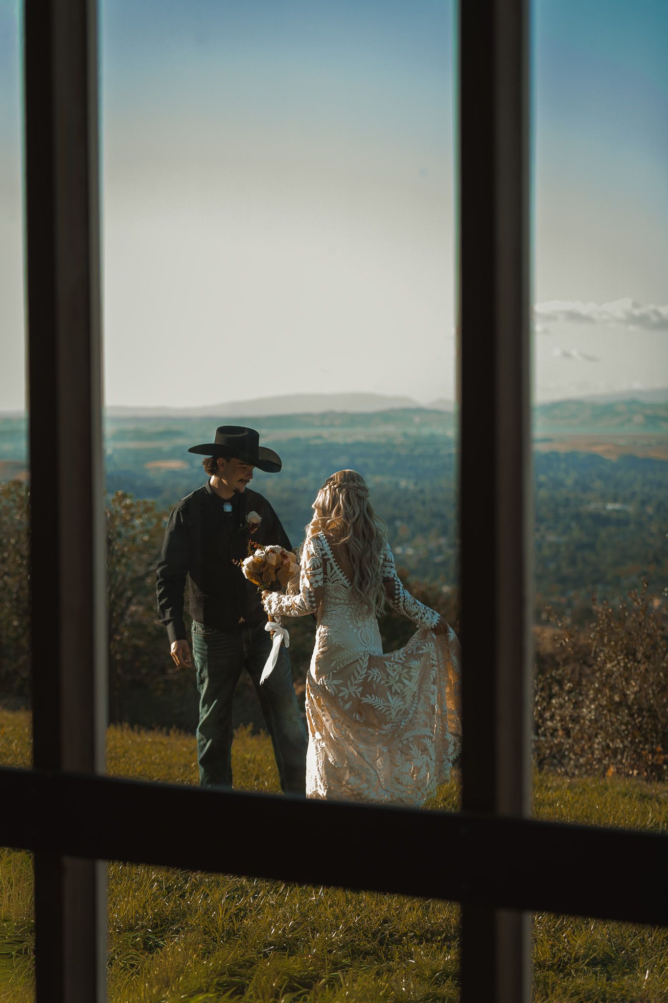 Wedding couple framed through window with scenic mountain landscape backdrop.