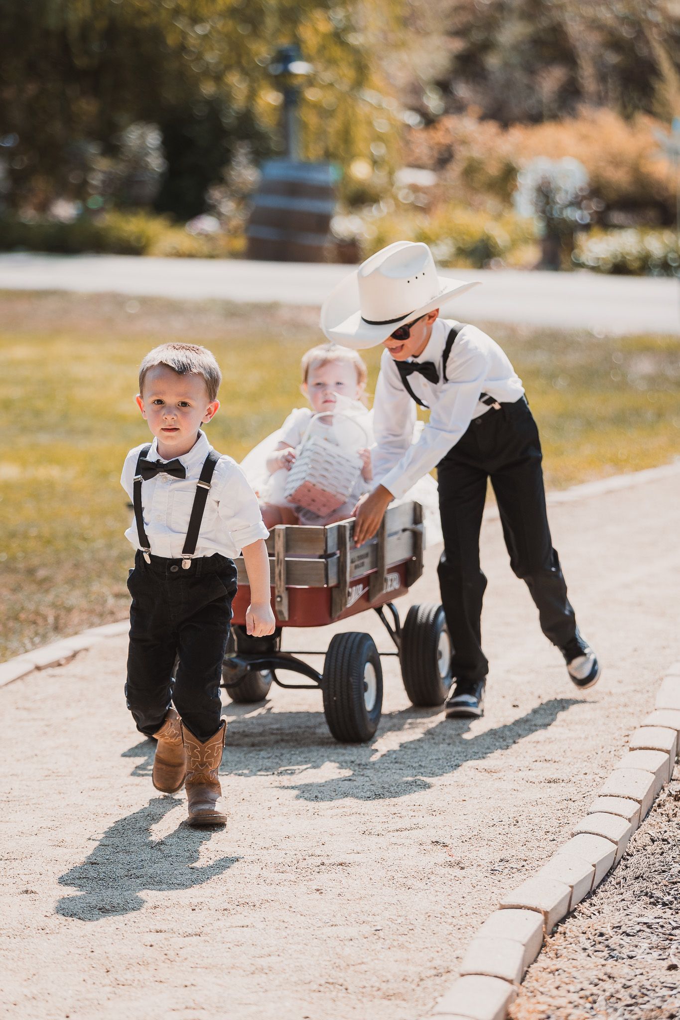 Young ring bearers in suspenders and bowties play on a path outdoors.