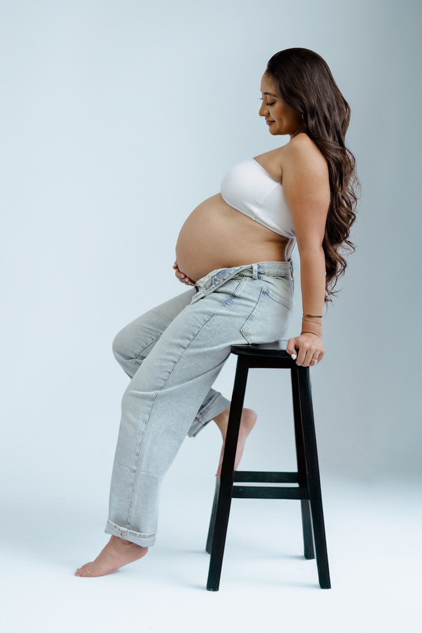 Person in light jeans sits sideways on a black stool against a light gray backdrop.