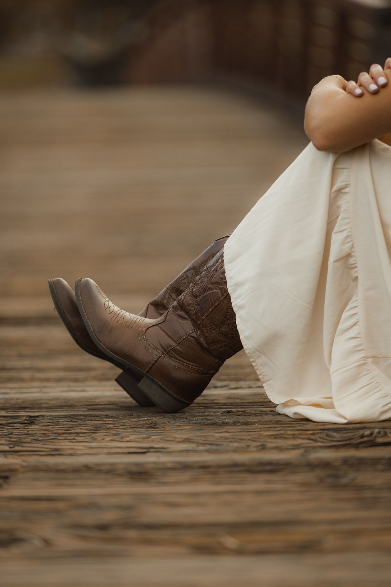 A pair of brown leather cowboy boots peeks out from under the hem of a flowing cream-colored dress on a wooden floor.