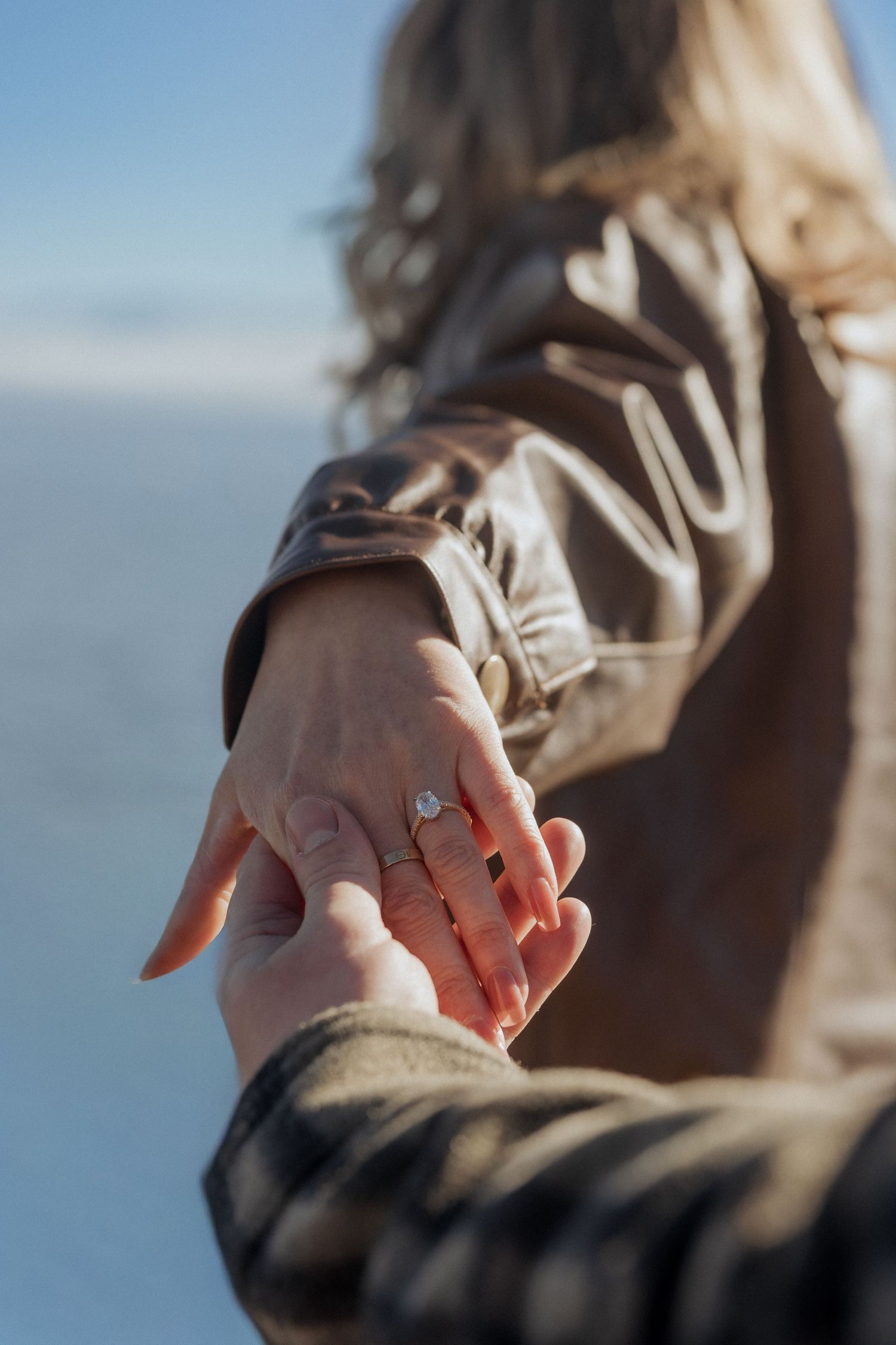 Close-up of a woman wearing an engagement ring while holding hands outdoors near a bright, scenic horizon.