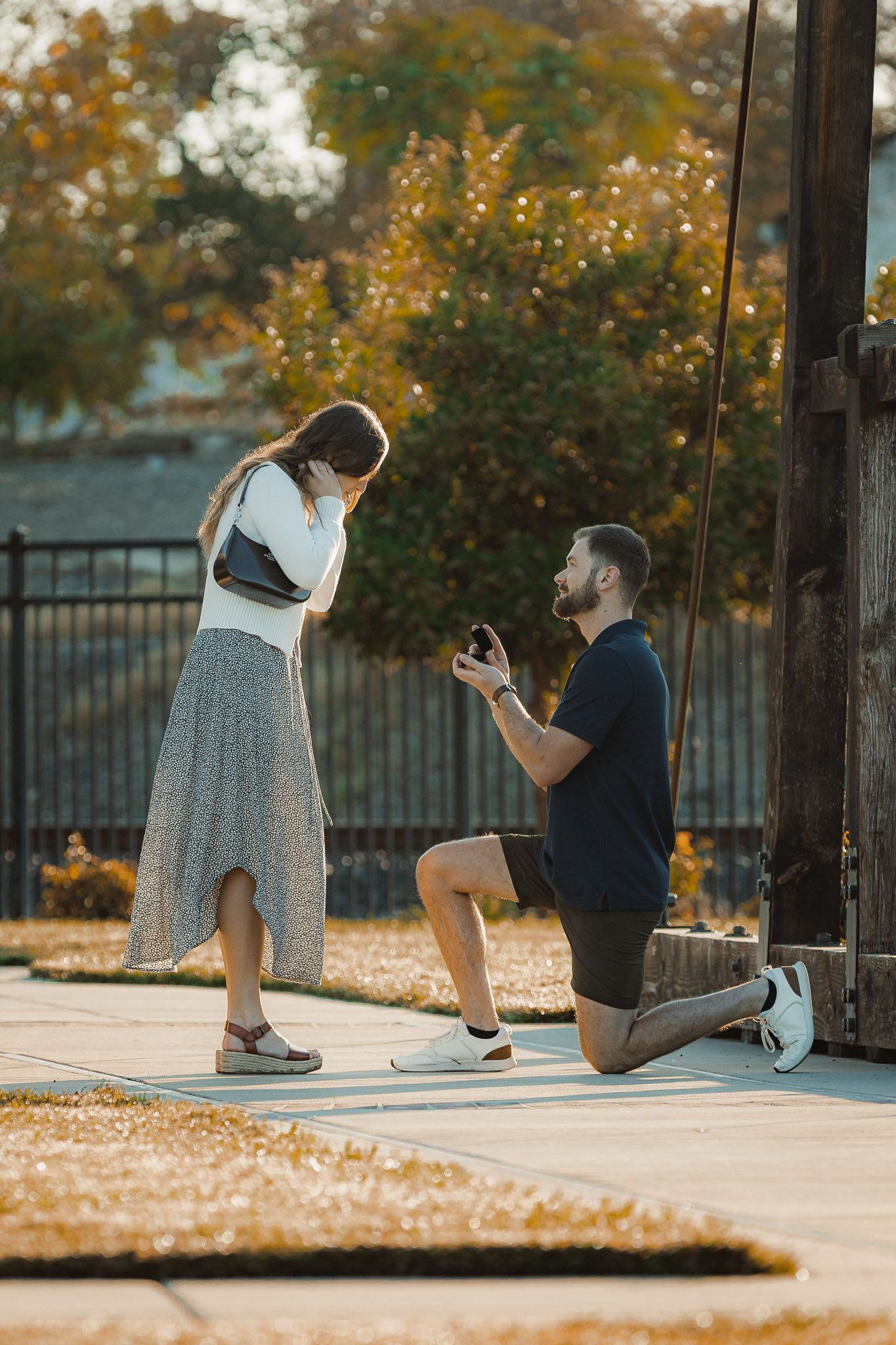 A couple sharing a romantic proposal moment in a park during golden hour as autumn leaves fall around them.