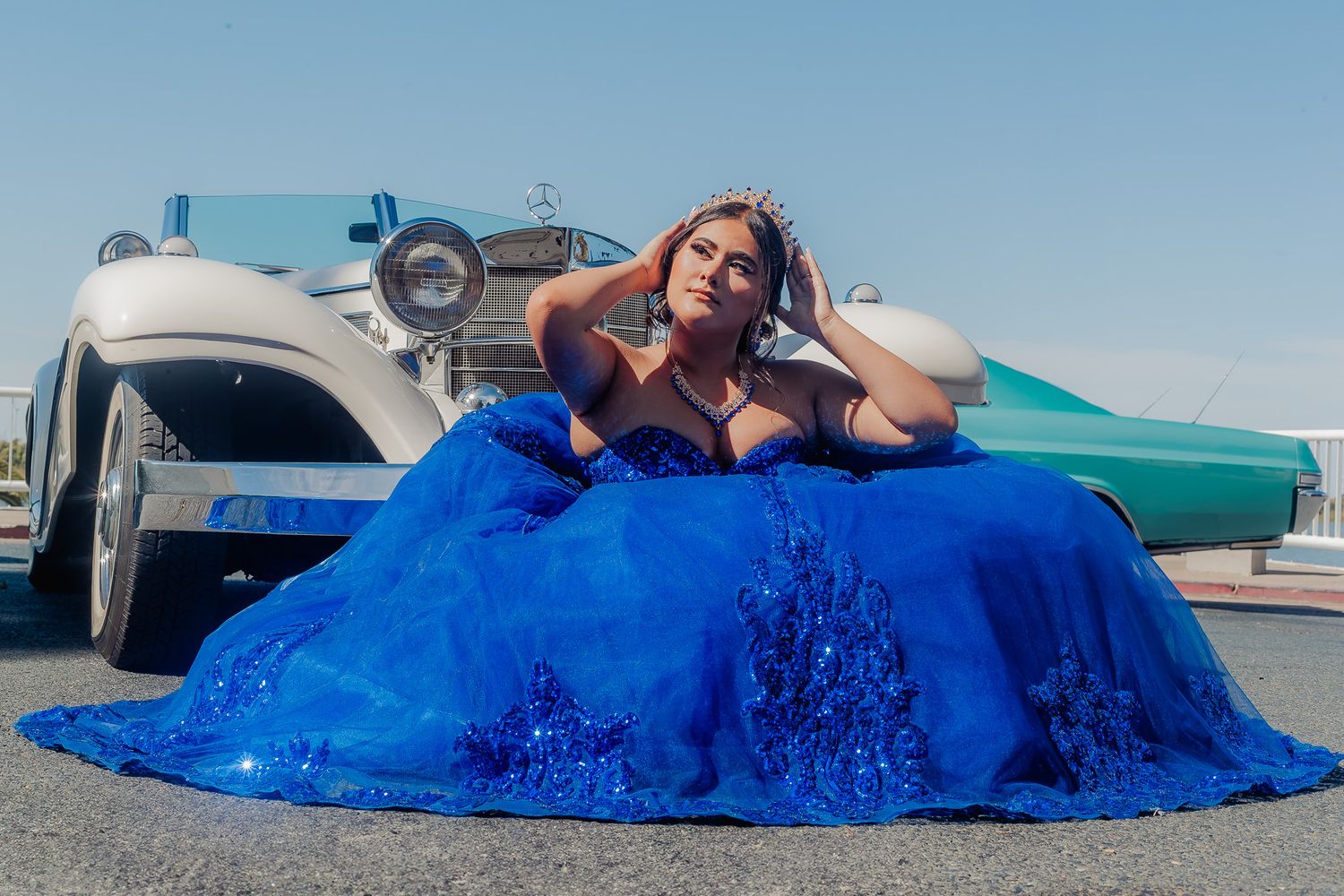 A person in a vibrant royal blue ballgown poses dramatically on the ground next to a vintage car.