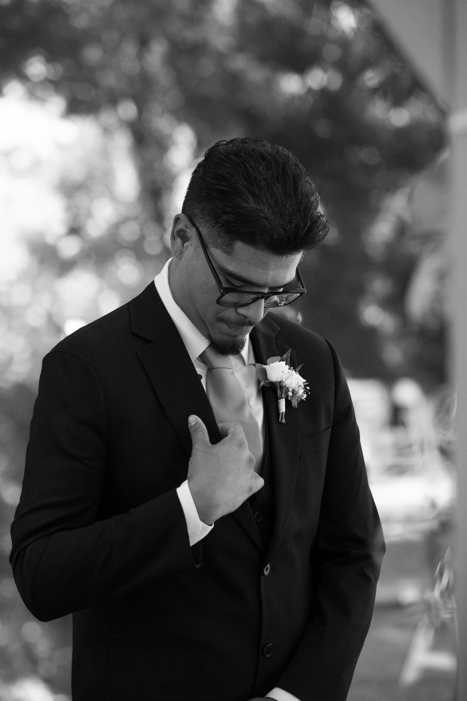 Black and white photo of a groom in a suit adjusting his tie with a boutonniere, looking down emotionally.