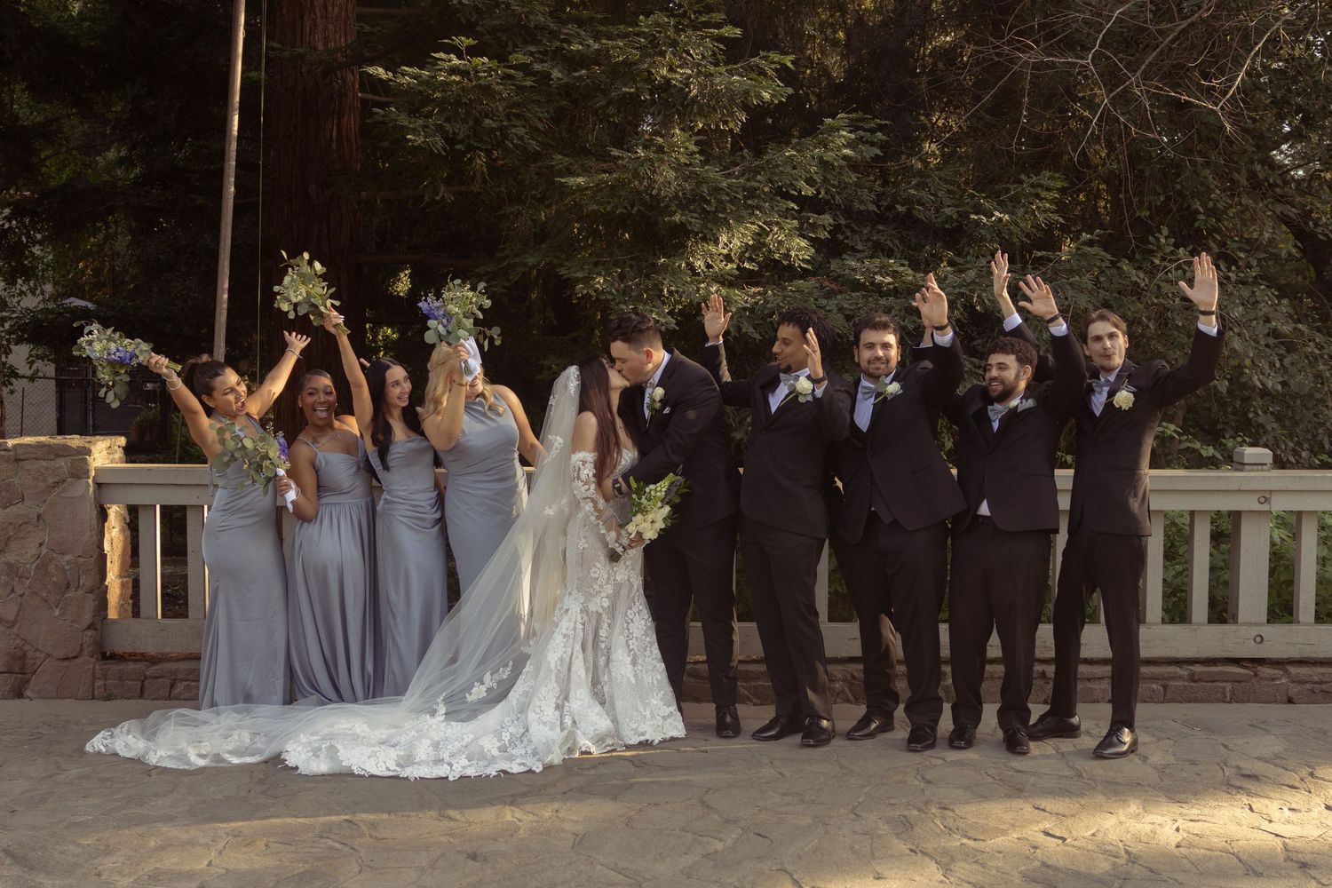 Wedding party poses outdoors, bridesmaids in blue-gray gowns and groomsmen in black suits, all cheering with arms raised.