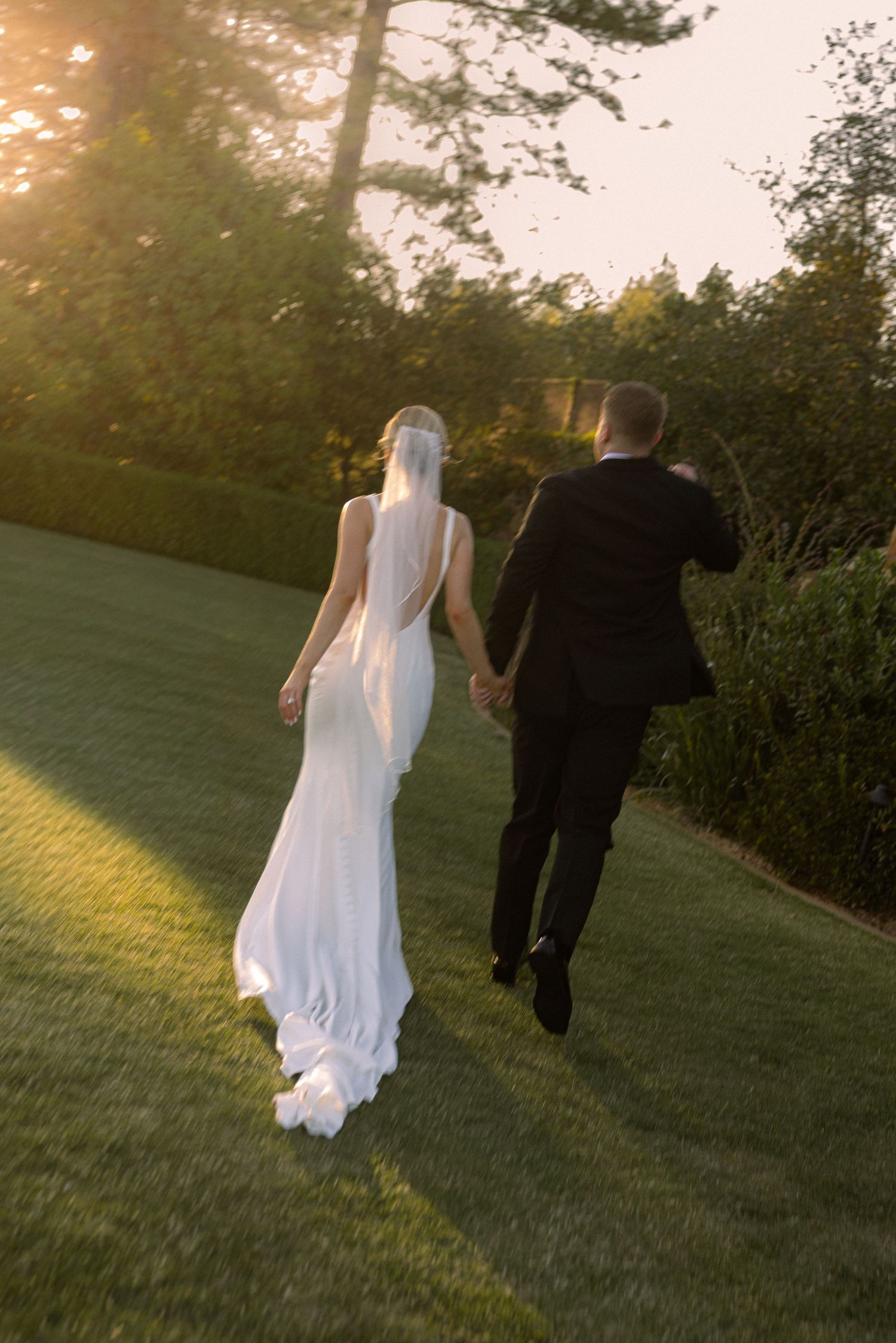 Bride and groom walking hand in hand across lush green lawn at sunset, bride in elegant white gown with veil.