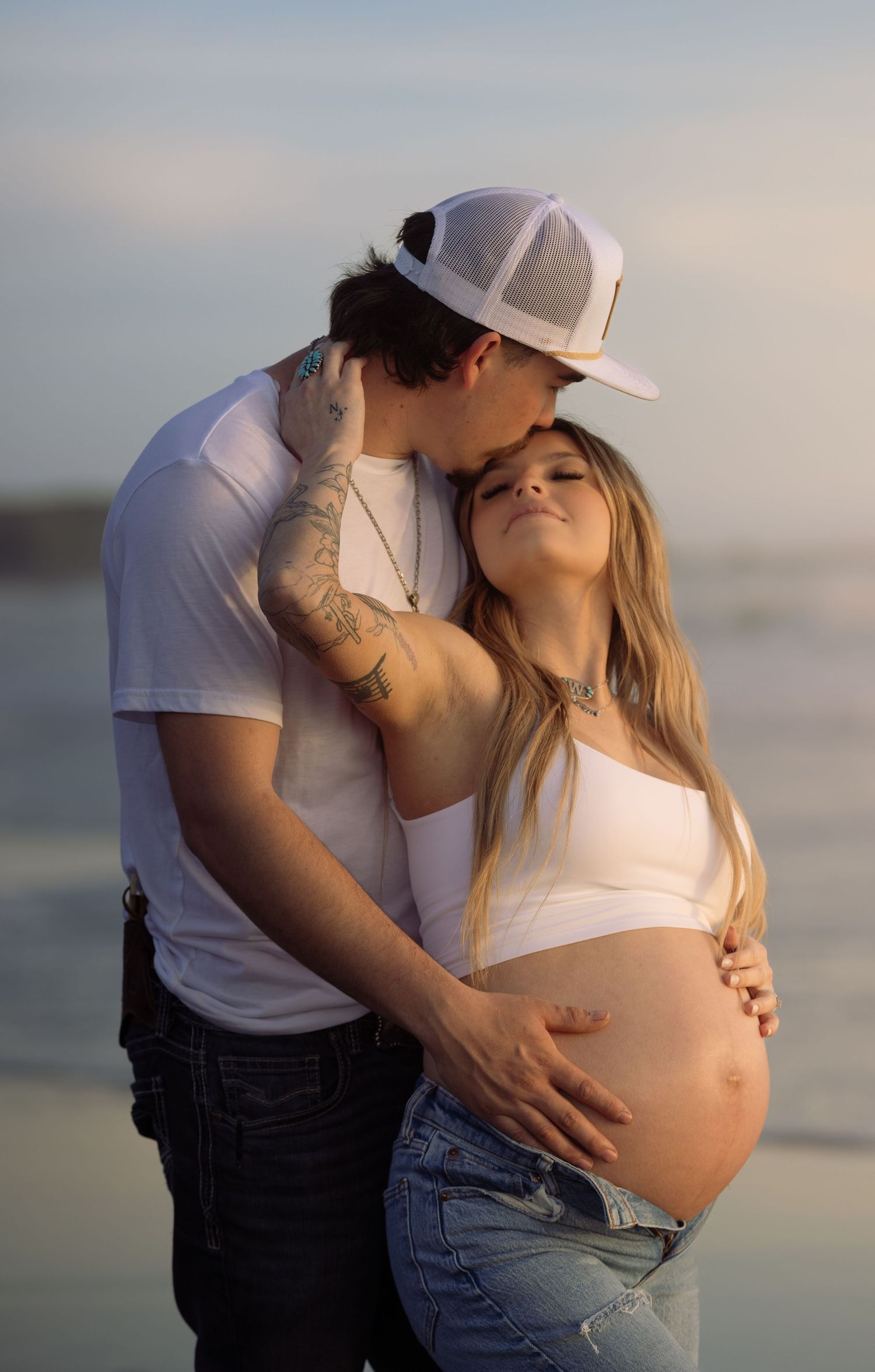 A couple shares a tender moment on the beach, with the man kissing his pregnant partner's head while cradling her baby bump.