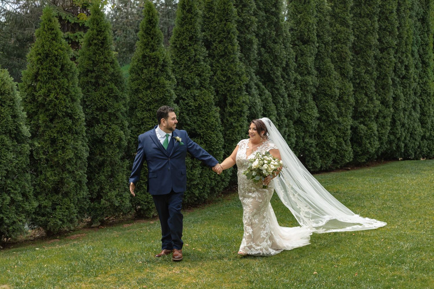 Bride and groom hold hands walking on green grass lawn with tall evergreen hedges backdrop at outdoor wedding.