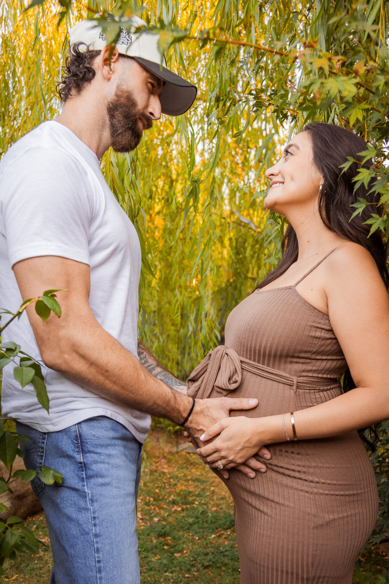 Intimate maternity photo shoot outdoors with couple embracing under willow trees in autumn setting.