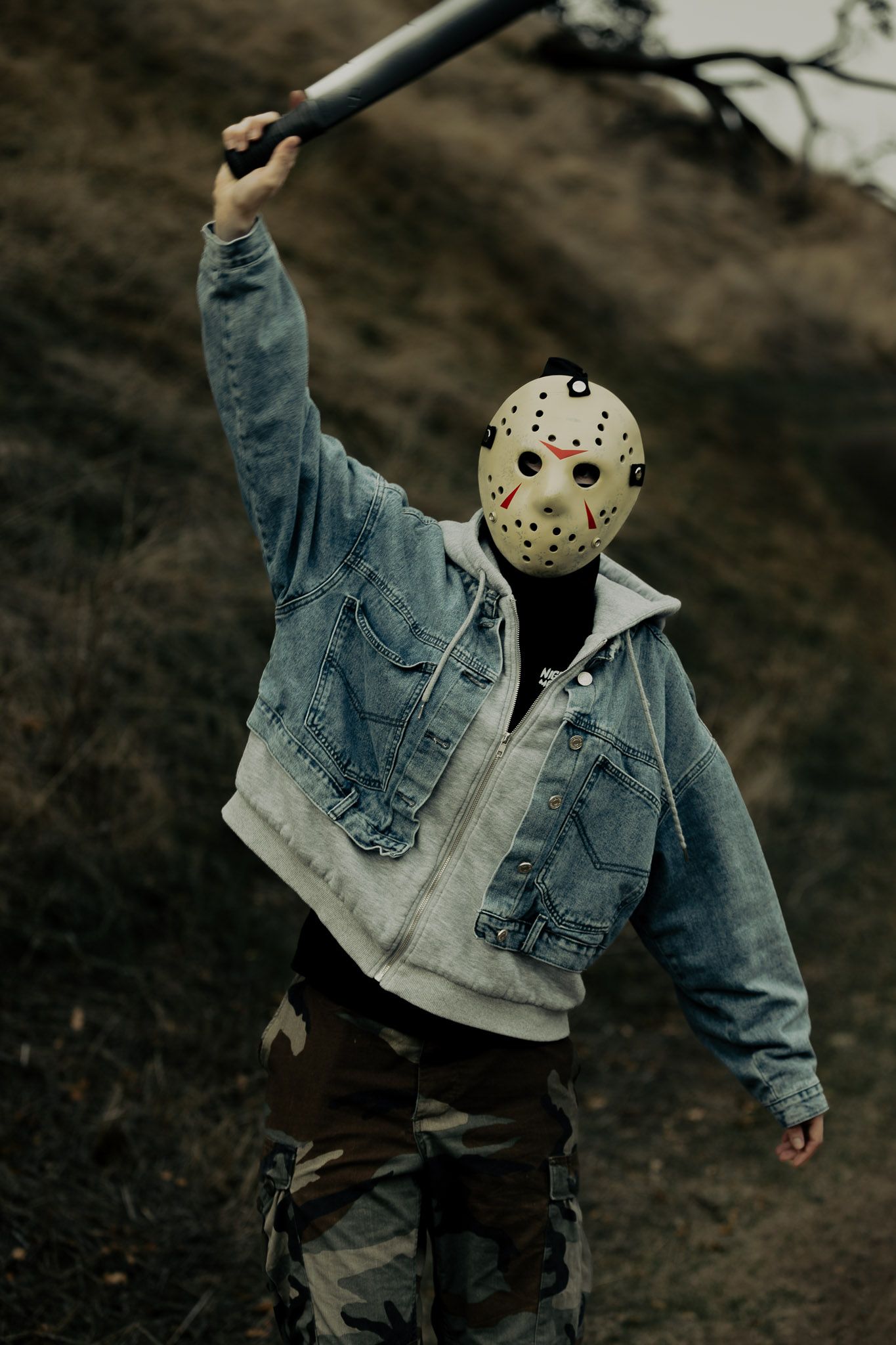 Figure in hockey mask and denim jacket holds up a weapon while posing in moody outdoor setting.
